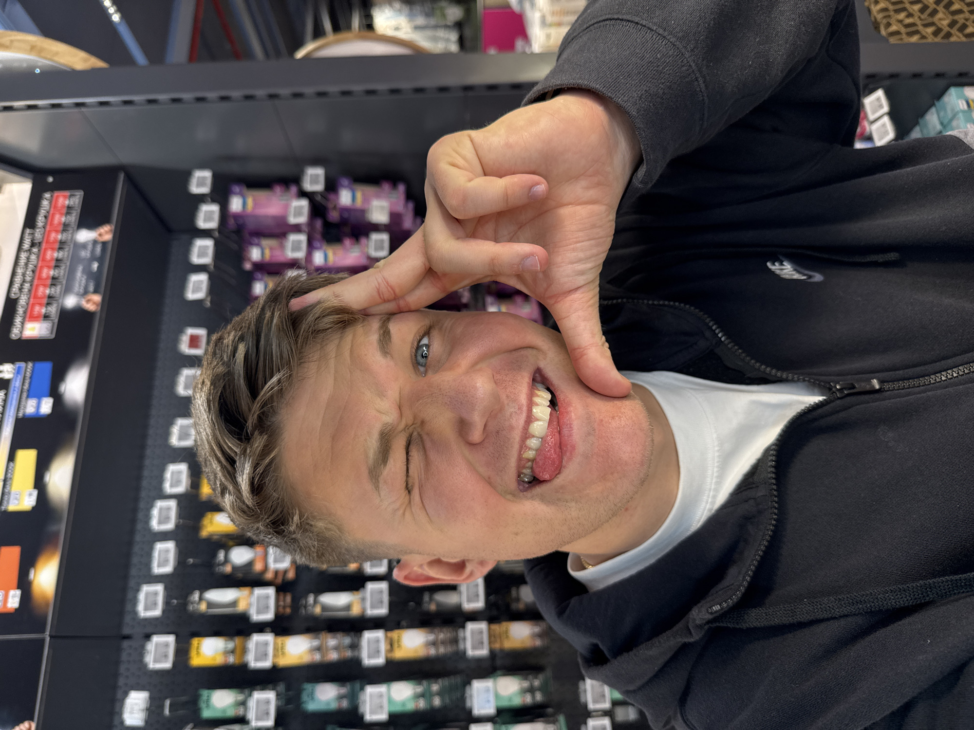 A man is posing in what appears to be a retail environment. He is wearing a black zip-up hoodie over a white t-shirt. He is making a silly face, squinting one eye and sticking his tongue out. His left hand is raised near his forehead, forming a shape. Behind him, there are shelves stocked with items, suggesting this is a store. The items on the shelves are too blurred to identify. The context of Plovdiv, Bulgaria is not visually apparent in the image.