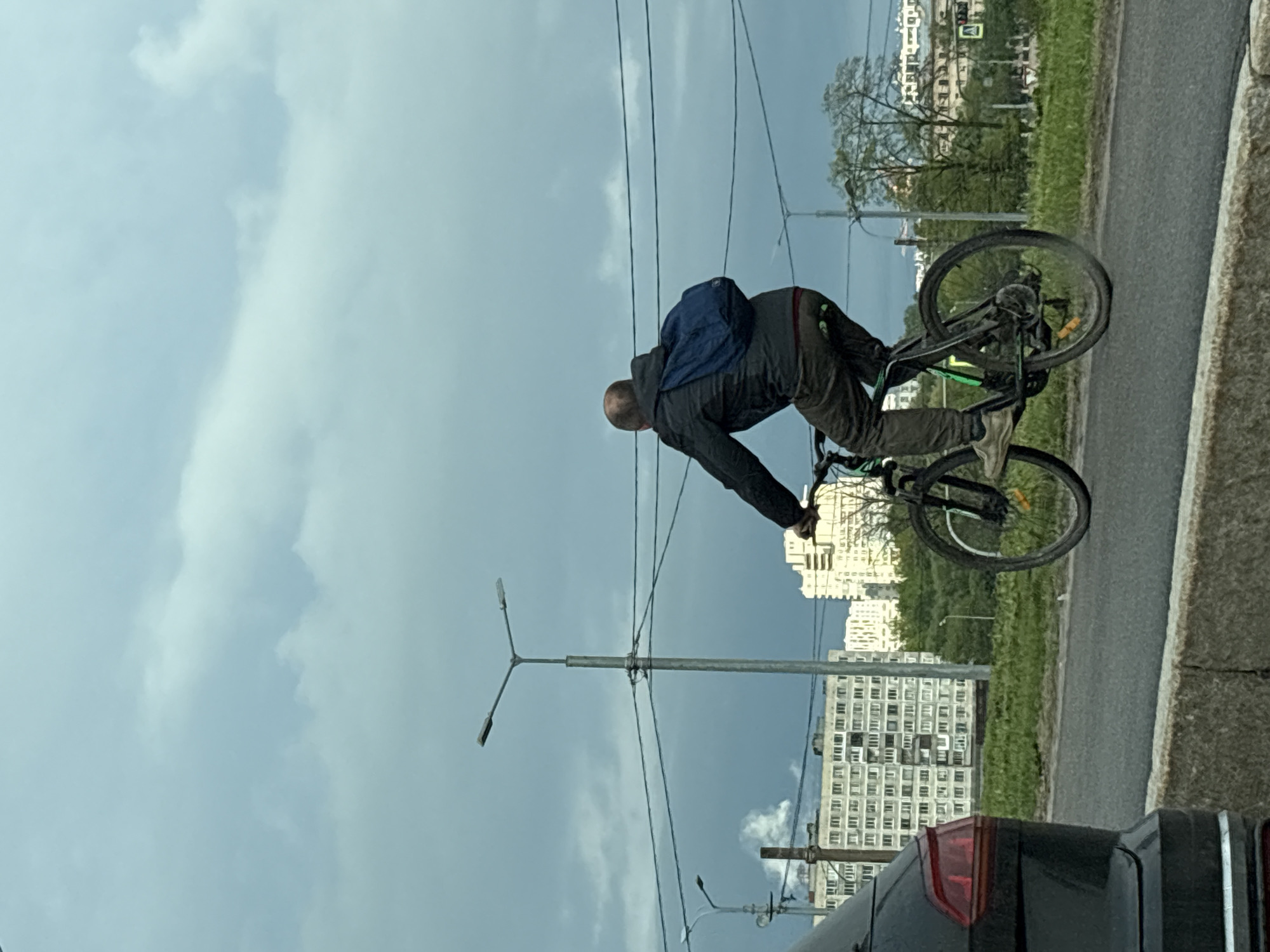 The image shows a man riding a bicycle on a street in Saint Petersburg, Russia. The man is wearing a dark jacket, pants, and a blue backpack. He is hunched over the handlebars of the bicycle. The bicycle appears to be black and green. Behind the cyclist, there is a gray road and a grassy area. Behind that is a tall, light-colored apartment building. In the foreground is a small portion of the tail light of a dark car. Above the cyclist, there are power lines and street lights. The sky is overcast with gray clouds.