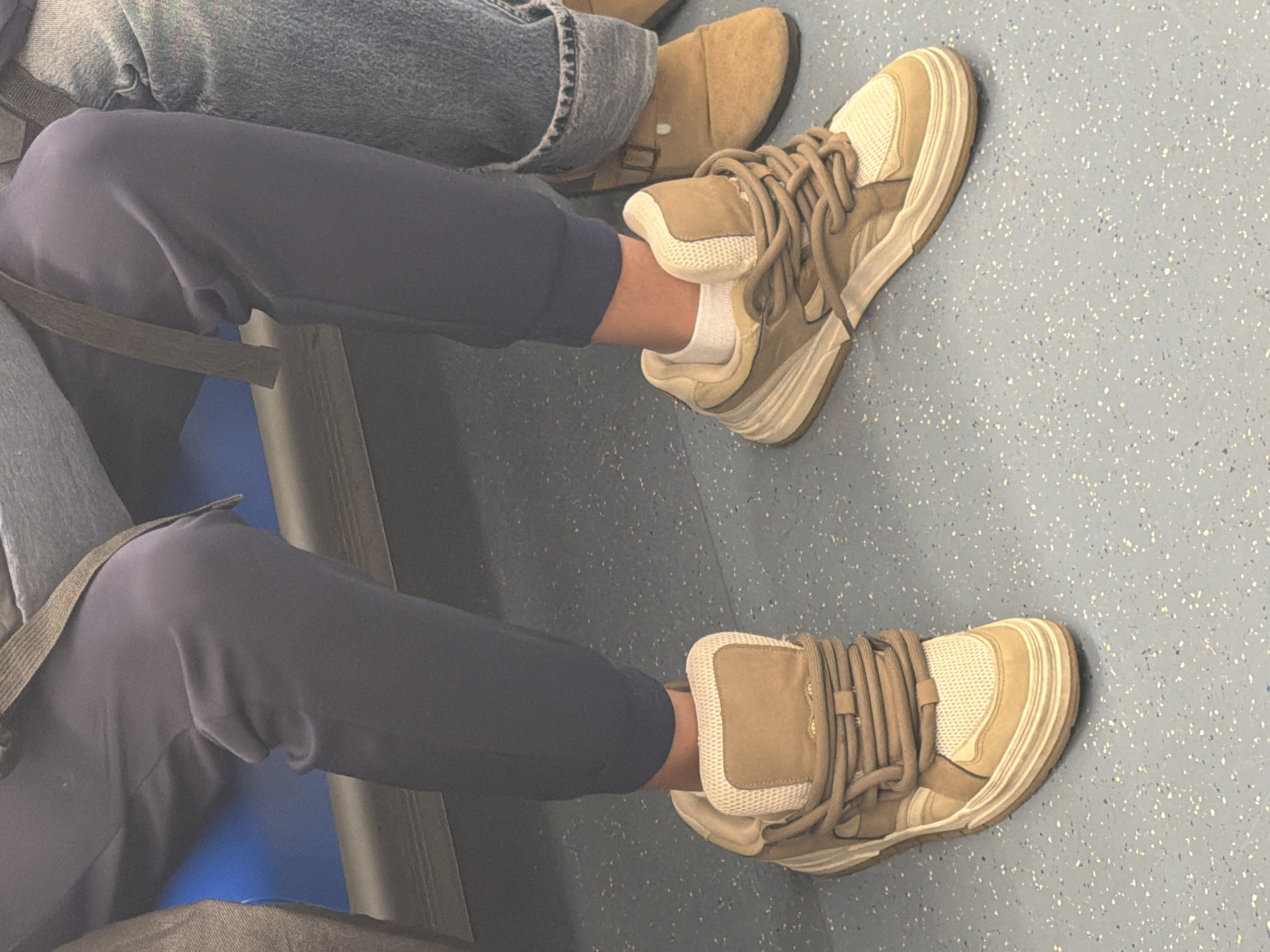 The image depicts two people seated, likely on public transportation. The person in the foreground wears dark blue sweatpants and off-white sneakers with prominent tan laces. White socks are visible. To the side, the second person wears blue jeans and brown suede shoes with a buckle. The flooring appears to be speckled blue. A portion of a seat and a seatbelt are also visible. The location is Unknown, Unknown.