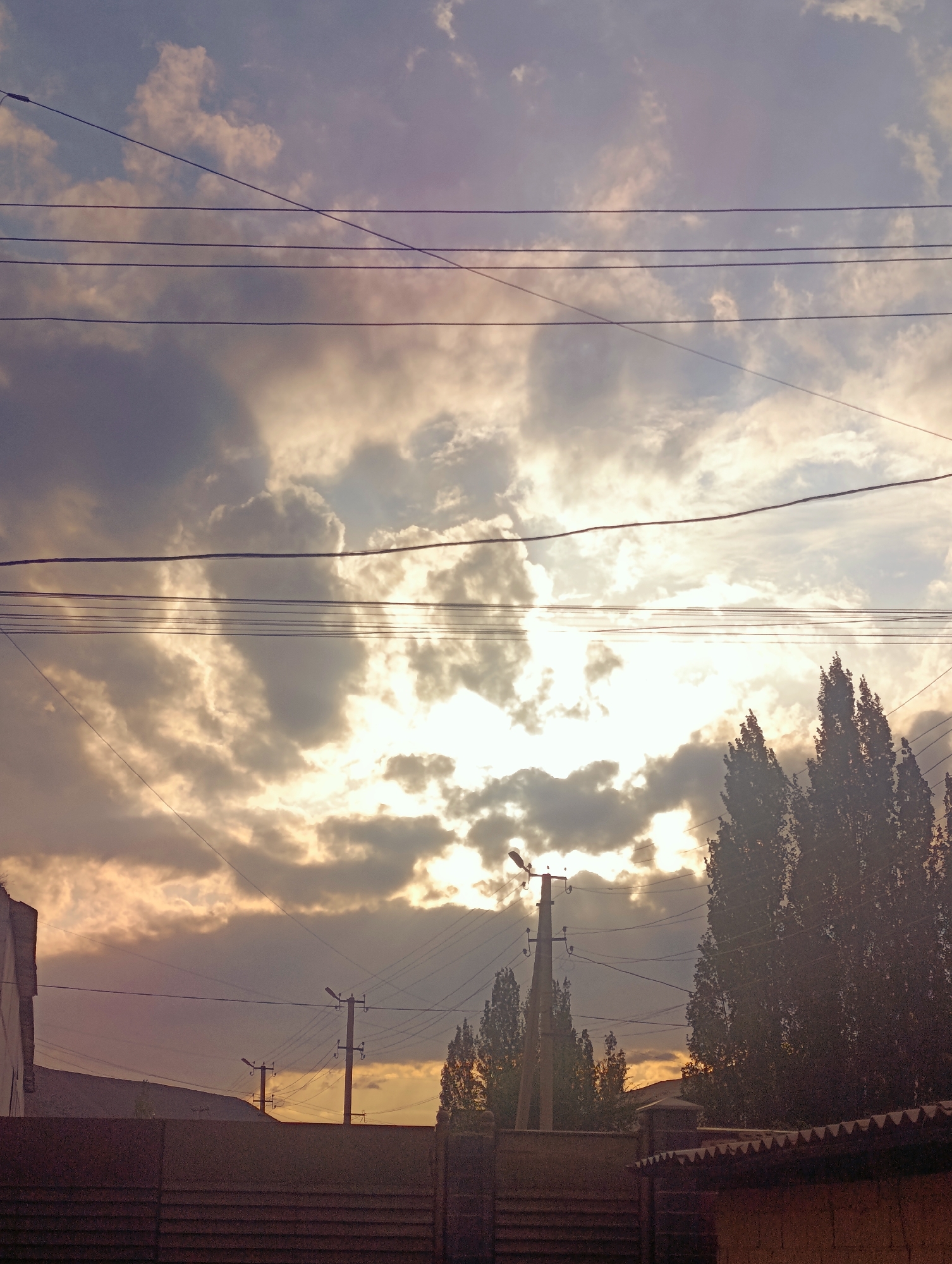 The image depicts a view of the sky above Naryn City, Kyrgyzstan, with several utility poles and power lines silhouetted against the clouds. The sun is partially obscured by the clouds, casting a warm, golden light across the scene. The clouds themselves are a mix of dark and light grays and whites, suggesting an overcast or partly cloudy day. In the foreground, a dark fence runs horizontally across the bottom of the frame, and to the right, the tops of tall, dark trees are visible. A street lamp is attached to one of the utility poles. The overall impression is one of a somewhat urban, possibly residential, area at dusk or during a dramatic weather event.