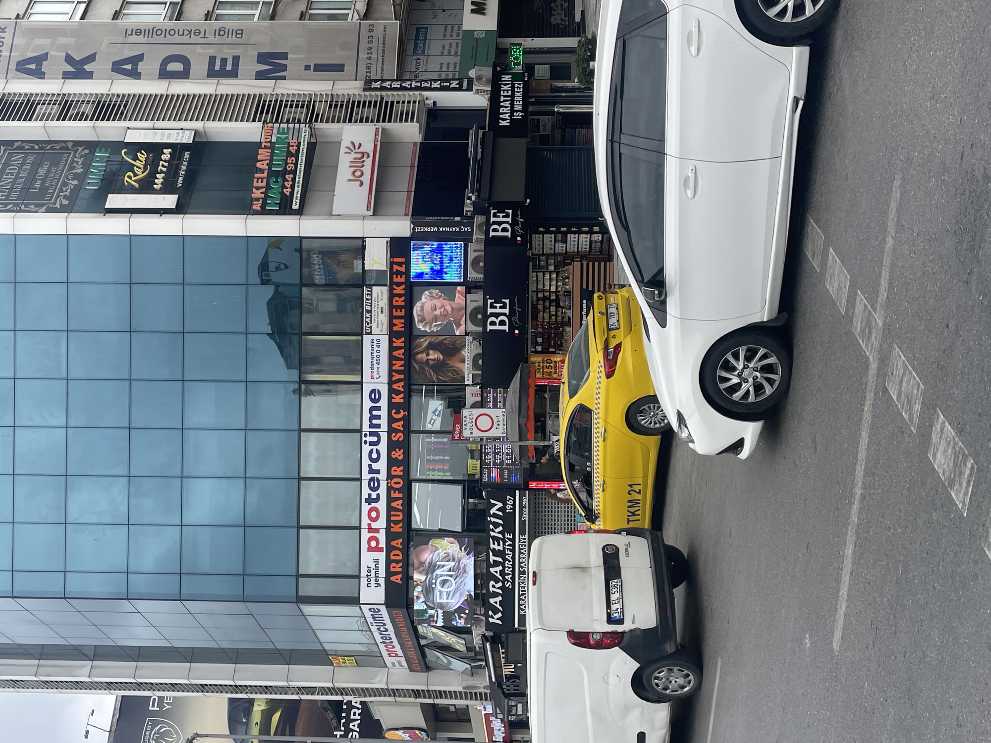 The image depicts a street scene in Kadıköy, Turkey, characterized by a multi-story modern building with extensive glass facades. Numerous businesses are advertised on the building's exterior, including "ARDA KUAFÖR & SAÇ KAYNAK MERKEZİ," "KARATEKİN SARRAFIYE," and "AKADEMİ" with the text "Bilgi Teknolojileri" beneath it. Several cars, including a white van and a yellow taxi, are present on the road. The yellow taxi has a license plate visible that reads "TKM 21." A white sedan is also prominently featured in the foreground. The environment suggests a commercial area with ongoing traffic. The lighting indicates it is daytime, with overcast skies.