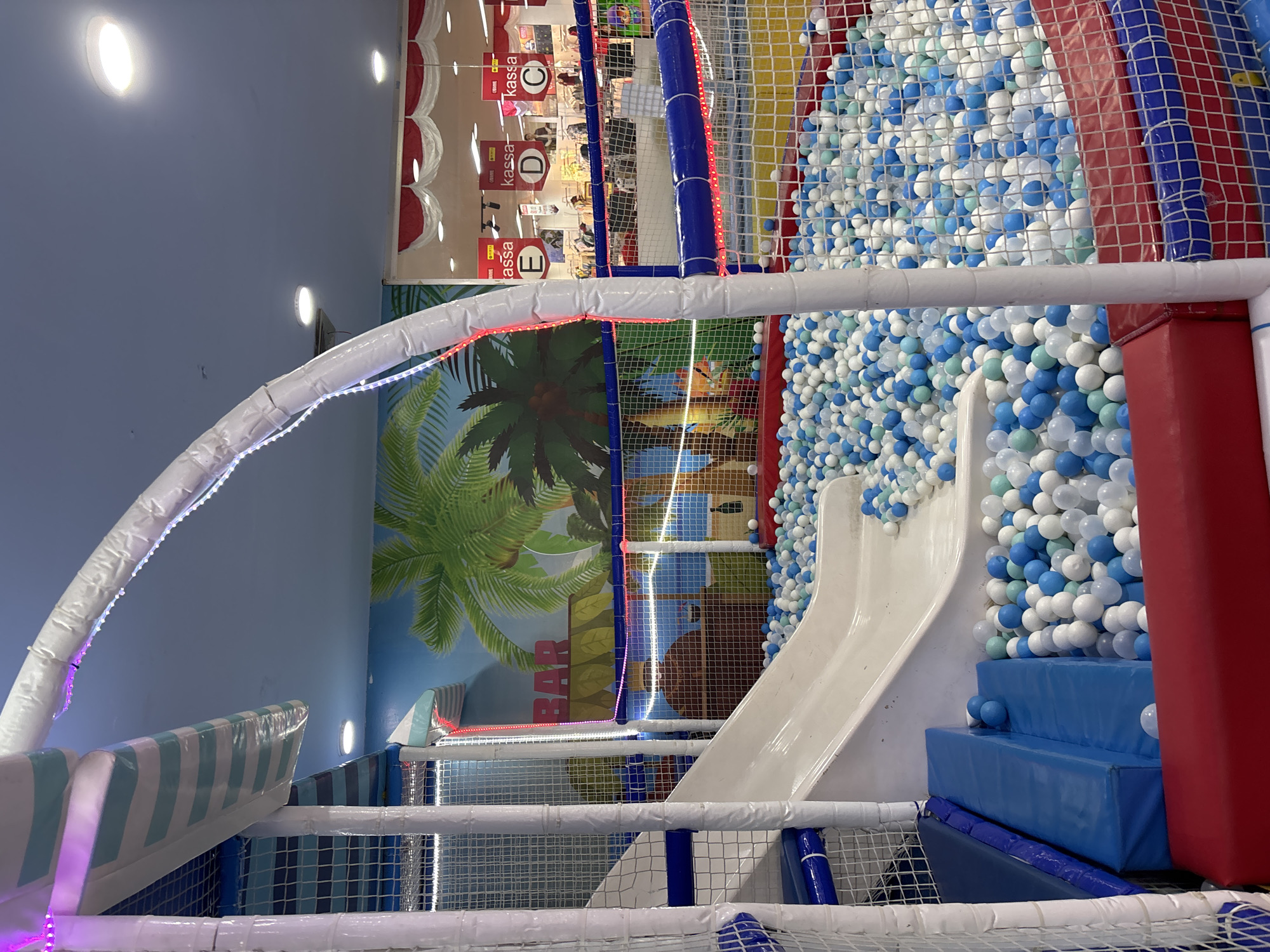 The image depicts an indoor playground, likely a ball pit area, with a white slide. The playground is enclosed by white and blue netting and features a mural of palm trees and a sign that reads "BAR." In the background, there appears to be a retail area with signs that say "kassa" and "Kass." The presence of "kassa" suggests this establishment is a retail store with a checkout area, possibly a supermarket or department store, located within the city of Liangjulang, Indonesia. The scene is brightly lit, and no people are clearly visible within the immediate playground area, though some figures can be seen in the background retail space.
