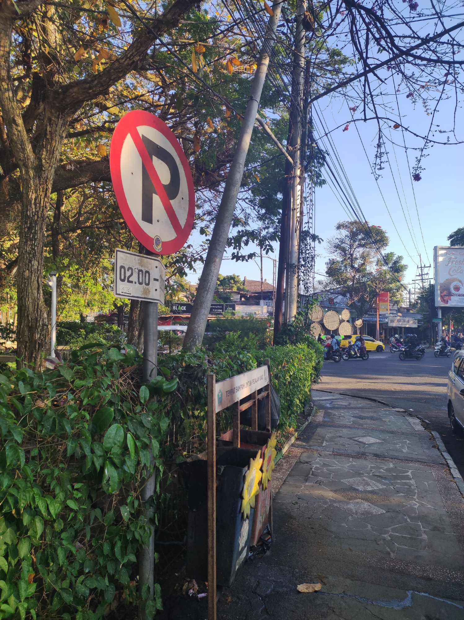 The image depicts a street scene in Malang, Indonesia, likely during daylight hours given the clear sky and sunlight. A prominent "No Parking" sign with a red circle and a diagonal red line over a black "P" is mounted on a pole. Below it, another sign indicates "02:00" and "SONER".
The foreground is dominated by lush green foliage, with a trash receptacle positioned behind the signs. To the right, a paved sidewalk with a pattern of irregular stone tiles runs alongside the road. The road itself is visible with a yellow car and several motorcycles with riders navigating the intersection. Buildings and trees are visible in the background, suggesting an urban environment. The overall impression is of a street corner with traffic and pedestrian infrastructure, marked by regulatory signage.