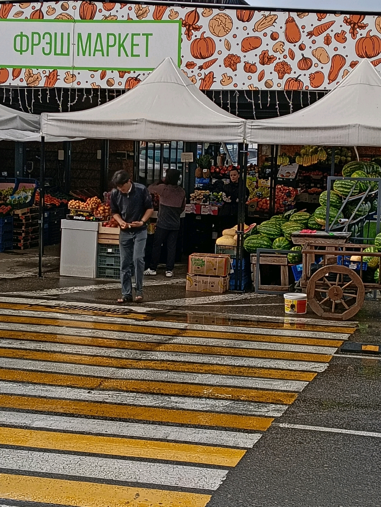 The image depicts an outdoor market scene, likely in Hrodna, Belarus, as indicated by the sign "ФРЭШ МАРКЕТ" (Fresh Market). The market stall features a variety of fruits and vegetables, including watermelons, pineapples, and what appear to be pomegranates and oranges. Several people are present: one person, dressed in a dark shirt and jeans, is standing on a crosswalk, seemingly examining something in their hands. Another person, wearing a dark top and pants, stands behind a stall. The ground is wet, suggesting recent rain, and the crosswalk stripes are prominent in the foreground. In the background, a colorful mural of produce decorates the market's facade. The overall atmosphere is that of a bustling, albeit damp, marketplace.