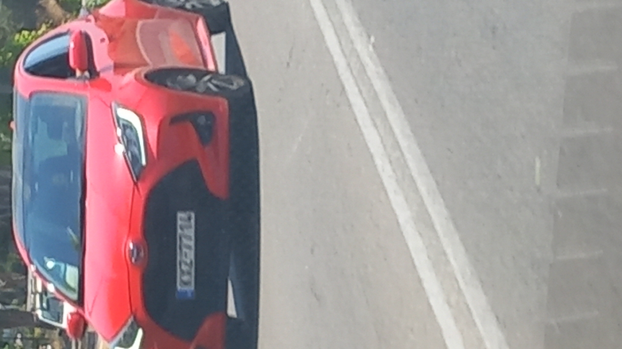 A red Toyota Yaris is driving on a paved road in an urban environment. The car has a black roof and a license plate that reads "KX2-7714". The road has two white lines painted on it. In the background, there is a white vehicle and some trees. The scene appears to be captured during daylight.