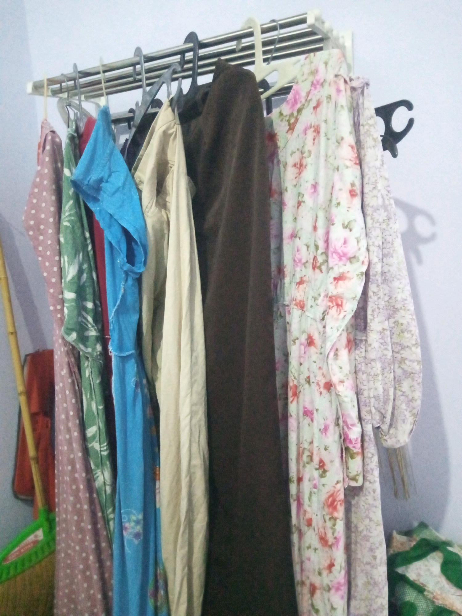 This image depicts a clothes rack with several garments hanging on it, likely in a domestic setting in Sumbergirang, Indonesia. The clothes rack is a white, multi-tiered metal unit mounted on a light blue wall. On the rack, from left to right, are a pink polka-dotted dress, a green floral-patterned dress, a red garment, a bright blue dress, a cream-colored garment, a dark brown garment, a light green floral-patterned dress, and a light purple floral-patterned dress. A black plastic hook is attached to the wall on the right side of the rack. To the left of the clothes rack, a portion of a broom with a green plastic head and a bamboo handle is visible, leaning against the wall. The scene appears to be indoors, possibly a laundry area or bedroom.