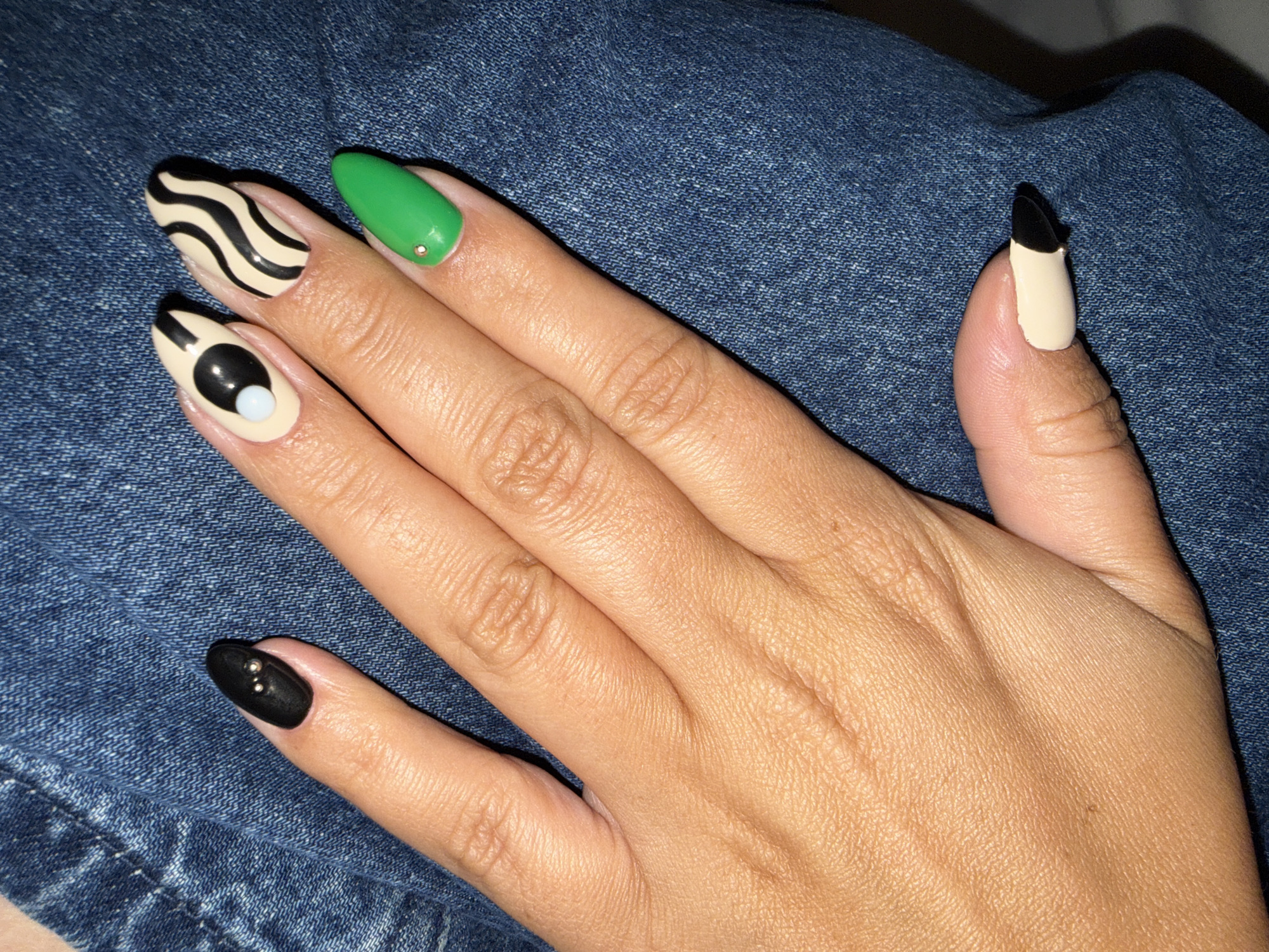 A close-up photograph shows a female hand with manicured nails resting on a blue denim surface. The fingernails are almond-shaped and feature different designs. The index finger nail is adorned with a zebra stripe pattern in black and beige. The middle finger nail is painted solid green with a small, circular, metallic-colored stud at the base. The ring finger nail has a beige base with a black dot and smaller white dot representing a yin-yang design. The little finger nail is painted solid black with two metallic studs placed vertically. The thumb nail has a beige base with the tip painted black. The denim background is slightly out of focus. No specific location information of North Kuta, Indonesia, is discernible from the image.
