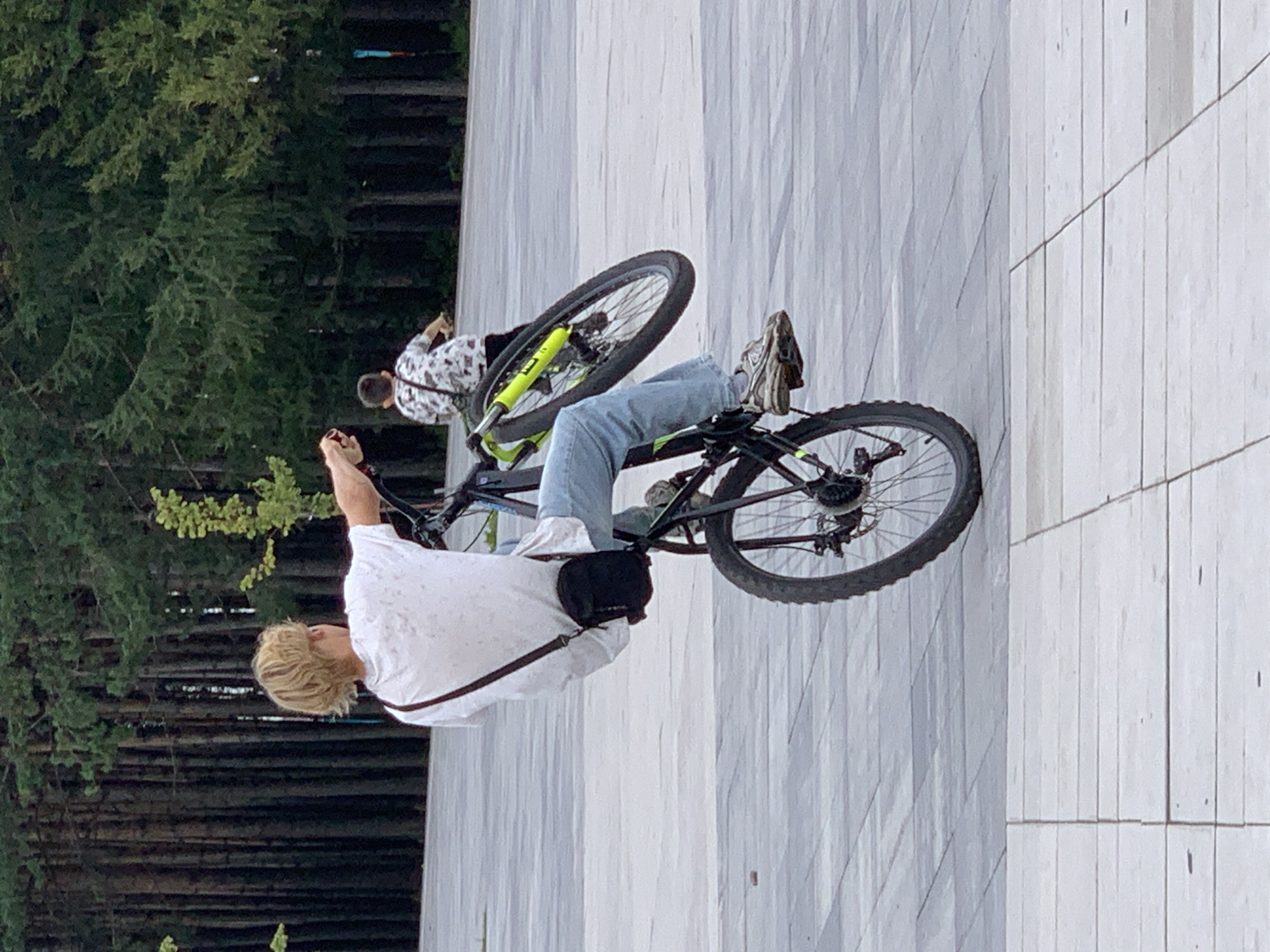 The image shows a young person with blonde hair performing a wheelie on a bicycle in an outdoor paved area. The person is wearing a white t-shirt, blue jeans, and athletic shoes, with a dark crossbody bag. The bicycle has a black frame and neon yellow accents on the front fork and wheels. In the background, another young person is riding a bicycle, and a dense forest of tall, dark trees lines the perimeter of the area. The ground is paved with large, light-colored square tiles. The lighting suggests it is daytime, possibly overcast.
The specific location of Prokopyevsk, Russia, cannot be determined from the visual information present in the image.