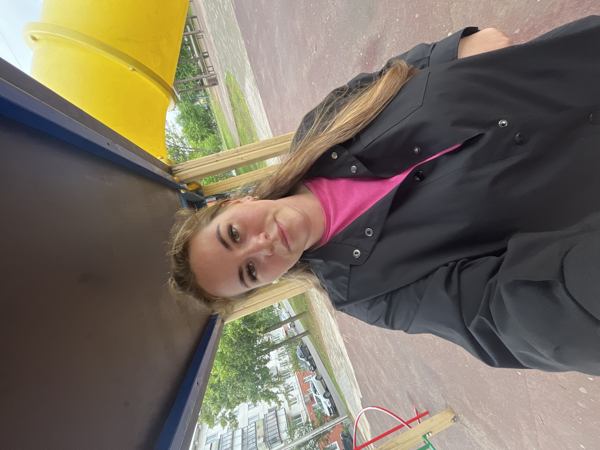 The media file is a slightly rotated, outdoor selfie featuring a young woman. She has long, blonde hair and is wearing a bright pink top under a black jacket. Her eyes are open and she is looking towards the camera with a neutral expression. The background is a playground with a large yellow slide, wooden beams, and a brown surface. Beyond the playground, there are trees and apartment buildings visible, suggesting an urban or suburban setting. The weather appears to be overcast, with no direct sunlight. The overall scene conveys a casual, everyday moment. There is no visible text in the image. The location context of Chaikovsky, Russia, is provided.