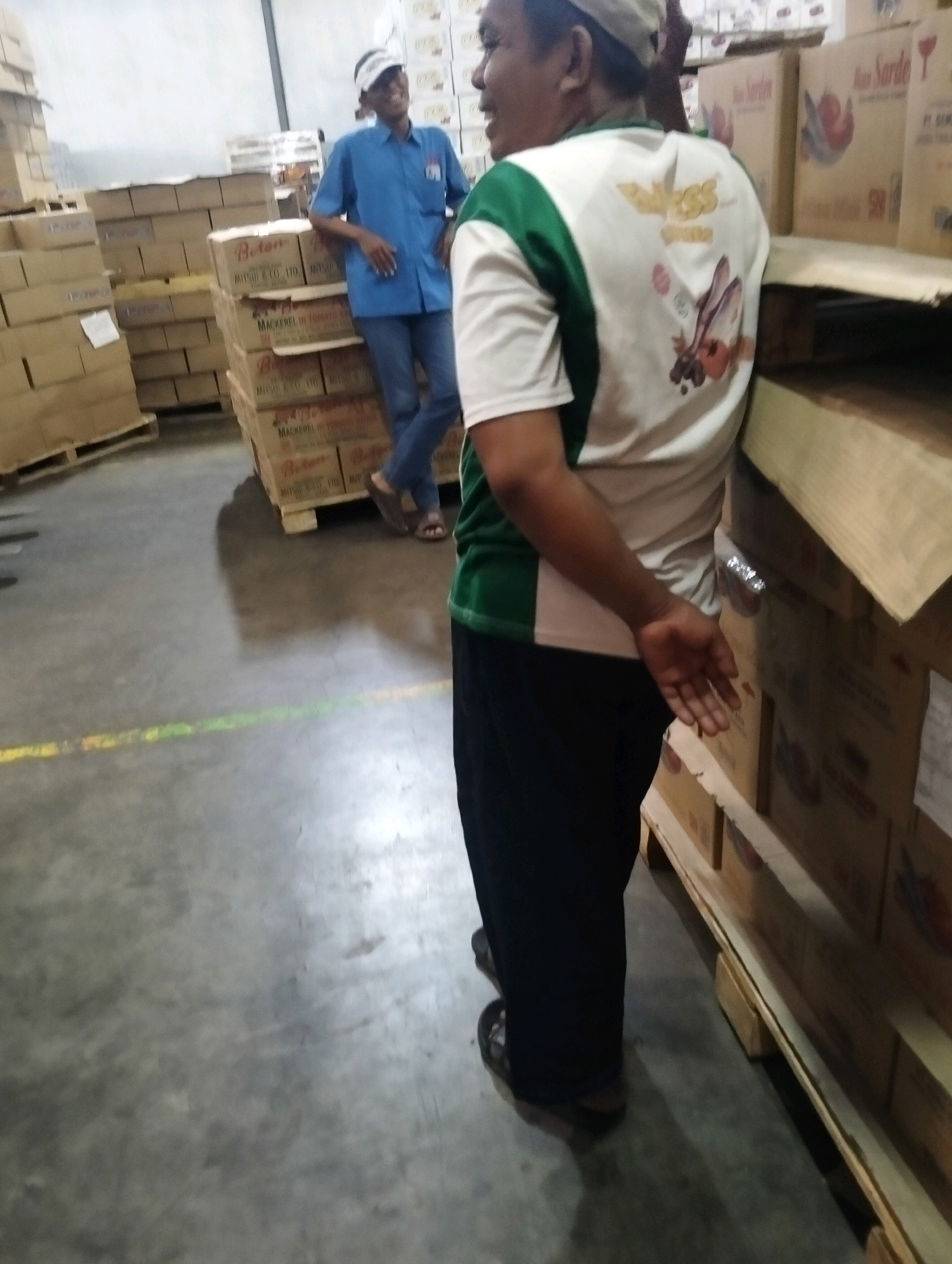 In Tanggulangin, Indonesia, a warehouse scene unfolds with two individuals amidst stacked boxes. The person in the foreground, viewed from behind, wears a white and green collared shirt with an emblem on the back and dark pants. Their right arm is bent, with their hand resting on a nearby stack of boxes. To the left, another individual, wearing a blue short-sleeved shirt and dark pants, stands with their hands on their hips, facing away from the camera and looking to their left. The environment is a concrete-floored warehouse, illuminated by overhead lighting, with numerous cardboard boxes organized on wooden pallets. Some boxes have visible labels, including "Boten" and "Mackerel in Tomato Sauce." The overall impression is one of an industrial storage facility.