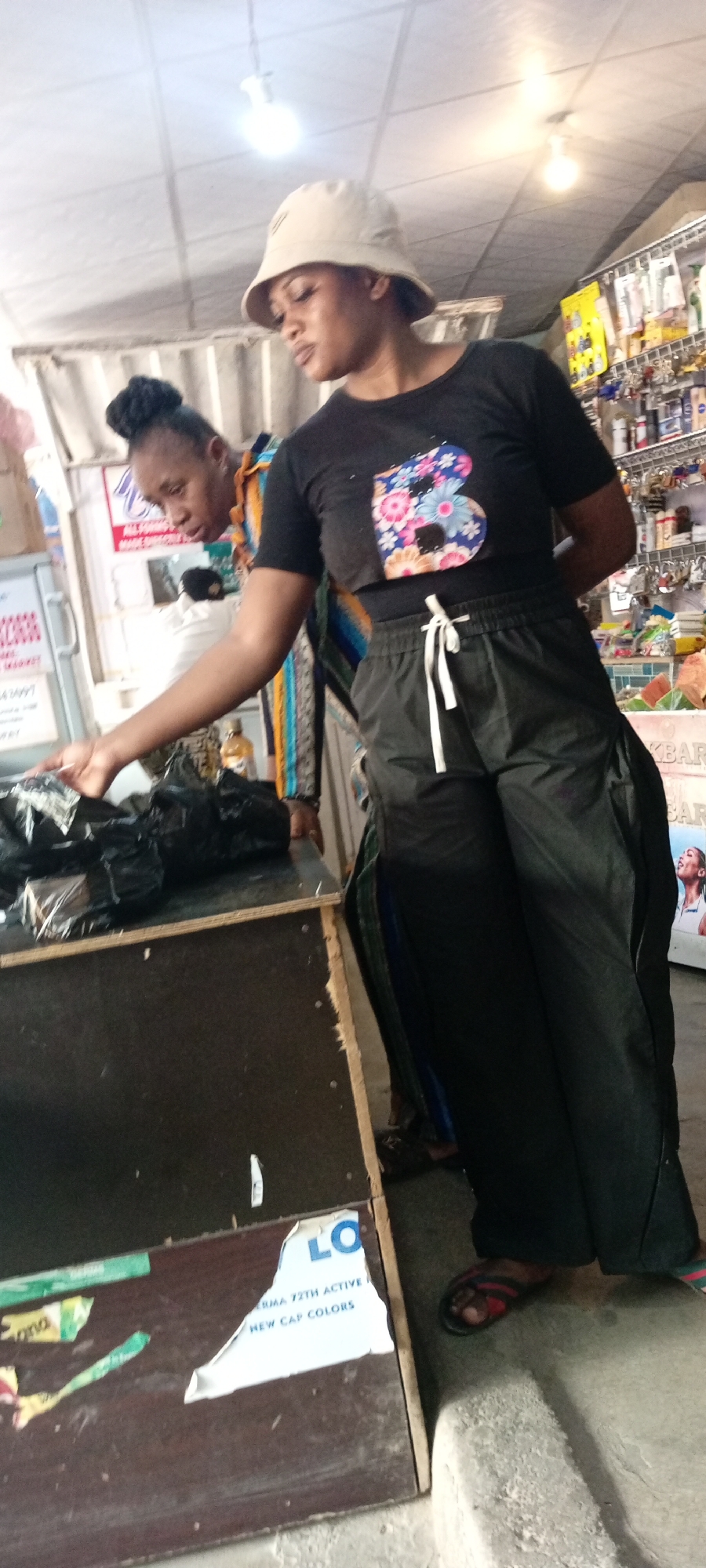The media file depicts a scene inside what appears to be a small shop or stall, possibly in Aviation Quarters, Nigeria. Two individuals are the primary subjects. The person in the foreground, wearing a light-colored bucket hat and a black crop top with a colorful floral "B" design, is looking downwards. They are also wearing black cargo pants with a white drawstring and flip-flops. Their posture suggests they are interacting with items on a dark counter in front of them.
Behind this person, another individual is visible, looking towards the counter as well. They have their hair styled in a bun and are wearing a white top underneath a brightly colored striped garment.
On the counter are several black plastic bags, suggesting merchandise or items being handled. The background reveals shelves stocked with various products, including what look like toiletries, cosmetics, or other small retail items. Above, ceiling lights illuminate the scene.
Visible text includes parts of a label on the counter which reads "LO" and "DERMA 72TH ACTIVE", and "NEW CAP COLORS". The overall impression is that of a retail environment, possibly a busy market stall, with a transaction or inventory check in progress. The lighting indicates it's likely daytime, and there are no clear indicators of weather conditions. The overall mood appears to be one of focused activity.