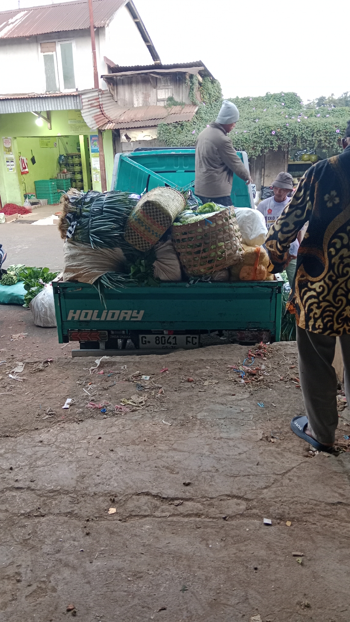 The image depicts a scene outside a building with a green facade, possibly a market or shop, in Klawen, Indonesia. A teal-colored truck is parked, laden with various produce, including bundles of green vegetables, woven baskets filled with produce, and large sacks. The truck has the word "HOLIDAY" written on its side and a license plate that reads "G. 8041 FC".
Two men are partially visible. One man, wearing a grey beanie and jacket, stands near the back of the truck, appearing to be arranging or inspecting the load. Another man, wearing a lighter-colored hat and a grey t-shirt, is visible behind him, also near the truck. A third person, wearing a patterned garment with gold and black designs, stands to the right, partially obscuring the view of the other individuals.
The ground in front of the truck is a rough, dirt-and-concrete surface littered with debris, including small colorful pieces of plastic and what appears to be dried plant matter. The building in the background features a corrugated metal roof, some wooden elements, and ivy growing up its side. The overall impression is a bustling market environment, likely in the morning or late afternoon given the lighting.