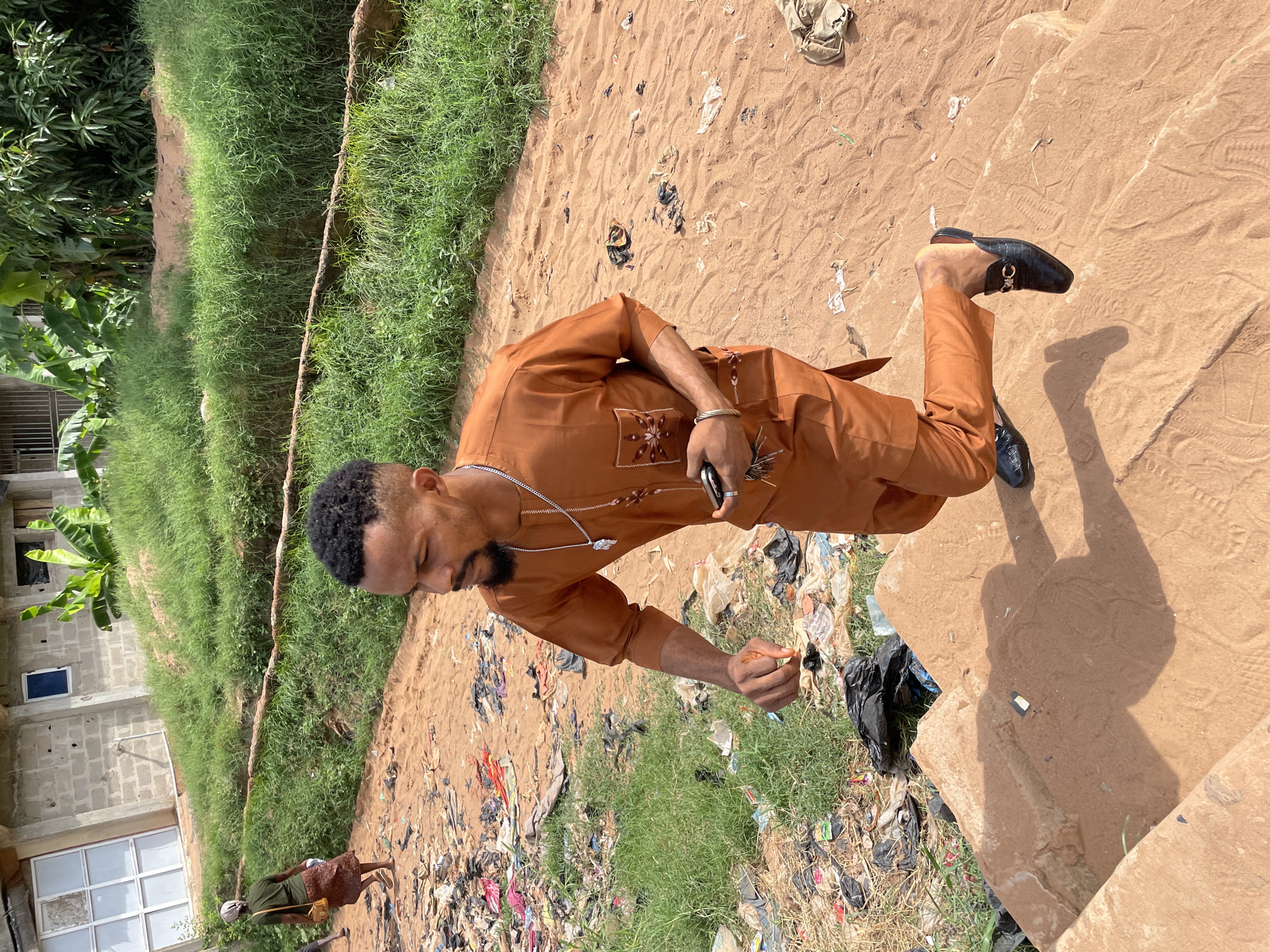 The image shows a man standing outdoors in Nkpor, Nigeria. He is wearing a brown traditional outfit and black loafers. He is holding a small object in his right hand, near a black plastic bag. The ground is a mix of sand, grass, and scattered trash. In the background there is vegetation. In the background, there is an unfinished building, a woman, and two children. There are several steps to the right and more sand with footprints on them.