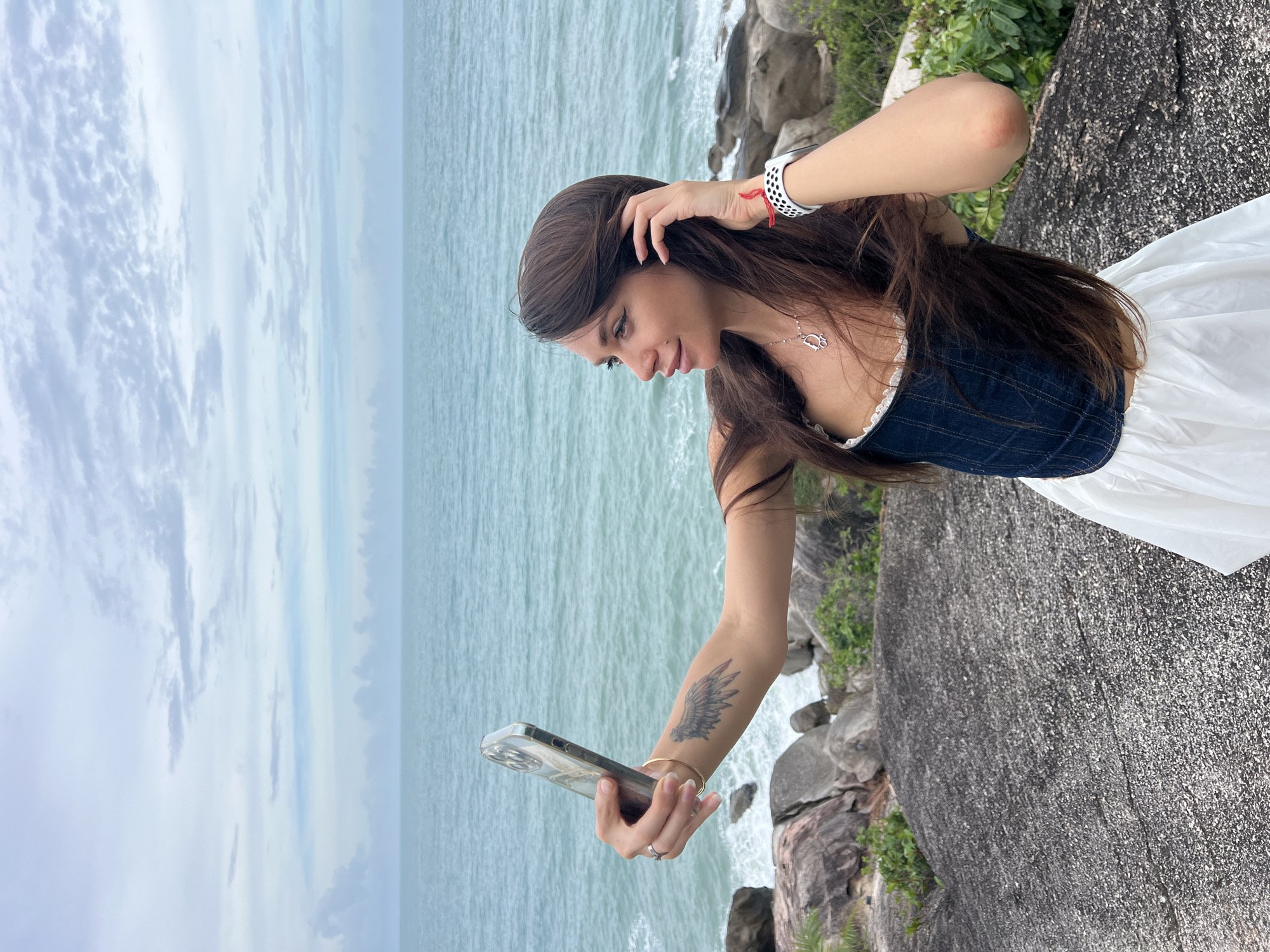 A young woman stands on a rocky outcrop in Baan Chaweng Noi, Thailand, holding a smartphone to take a selfie. The ocean stretches out behind her, with a cloudy sky above. She is wearing a dark top with white trim and a white skirt, and has dark hair. A tattoo is visible on her forearm. She is partially obscured by a large, dark rock. Vegetation grows nearby.