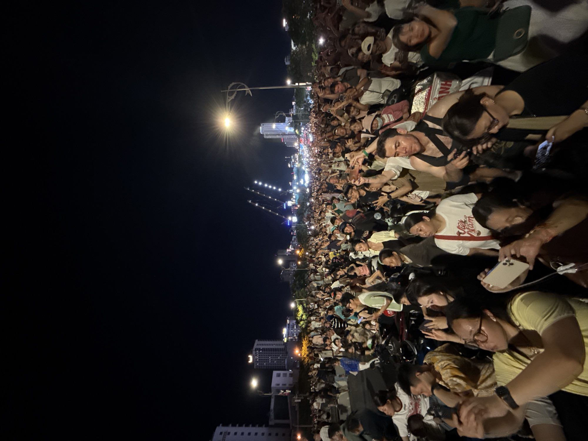 This image captures a dense nighttime gathering on a wide street or bridge in Da Nang, Vietnam, specifically identifiable by the illuminated Dragon Bridge (Cầu Rồng) in the background. Hundreds of people, predominantly of Asian descent, are tightly packed across the frame, extending into the distant background. Many individuals are standing or seated on numerous motorbikes that are also tightly packed together, indicating a public thoroughfare. A significant portion of the crowd is holding smartphones aloft, actively recording or photographing an event occurring towards the mid-left area, beyond the immediate foreground. People's gazes are largely directed towards a common point of interest in that direction.
Visible structures include multi-story buildings in the background, one on the left bearing partial "HOTEL" signage. Several streetlights illuminate the scene from above, with one on the right featuring a decorative, stylized fish or "S" motif on its pole. The Dragon Bridge in the distance is clearly discernible by its unique architectural profile and string lighting, characteristic of its design over the Han River. The sky above is dark, indicating night, while the urban environment is brightly lit by artificial sources. The overall scene suggests a large public event or a collective viewing of a spectacle.