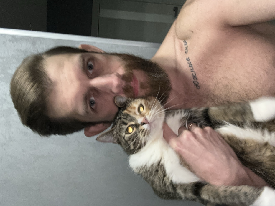 A selfie captures a shirtless man with brown hair and a beard holding a cat. The cat has striking yellow eyes and mixed brown, white, and grey fur. The man’s tattoo is visible on his shoulder, featuring Russian text. He is looking directly at the camera. A round object appears to be placed on the man’s forehead, near his hairline. The background is plain and slightly blurred.