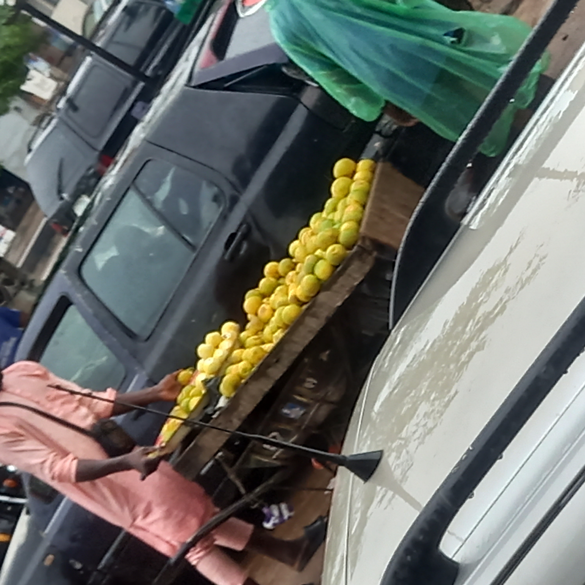 A medium shot shows a vendor in Sardauna, Nigeria, selling fruit from a table set up next to parked cars. The vendor, dressed in pink, is reaching for the fruit. The fruit is yellow and stacked on a wooden table. A person wearing a green raincoat stands near the fruit and a black car. Cars are parked along the roadside.