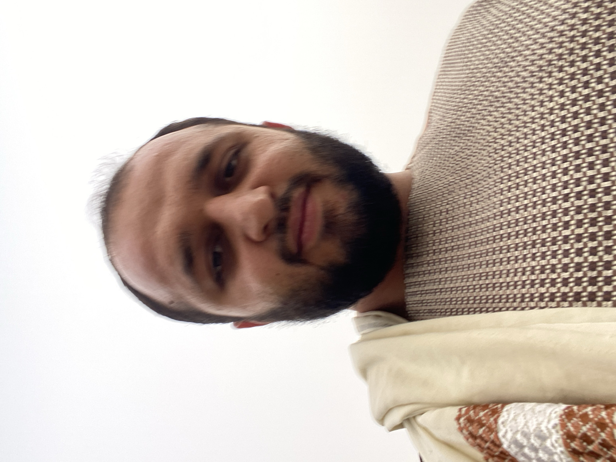 A man with a beard and receding hairline, wearing a patterned sweater and a cream-colored shawl with brown and white stripes, poses for a selfie. The background is a bright, overcast sky. The man's face is partially obscured by the angle of the photo and the shawl. The image appears to have been taken outdoors in Kandahar, Afghanistan.