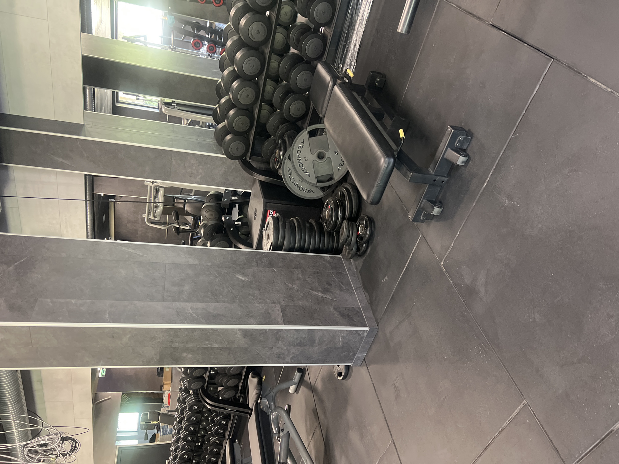 This image captures the interior of a gym, likely situated in Shumen, Bulgaria. The scene depicts an area dedicated to free weights and strength training, devoid of any individuals.
In the foreground, a black adjustable weight bench with yellow levers is positioned on dark gray rubber floor tiles. Behind the bench, a black metal rack holds multiple pairs of rubber-coated dumbbells, neatly arranged by ascending weight, with numbers such as 22, 24, 28, 32, 34, and 36 visible on their ends. To the left of the bench, a stack of circular Technogym branded weight plates, some clearly marked "20 kg," rests on the floor next to a black cuboid platform or box.
The gym features mirrored walls that extensively reflect the interior space, creating an illusion of greater depth and replicating equipment. Dark gray, marble-patterned tiled columns with white trim are integrated into the mirrored design. Above the mirrors and columns, the upper walls are painted in a light gray or beige tone. The ceiling reveals exposed spiral metallic ventilation ducts and bundles of white and black electrical wiring. A green "EXIT" sign is visible in a mirror reflection on the left side of the frame. The floor consists of large, dark gray rectangular rubber tiles with distinct grout lines. No people, actions, or direct interactions are visible within the frame.