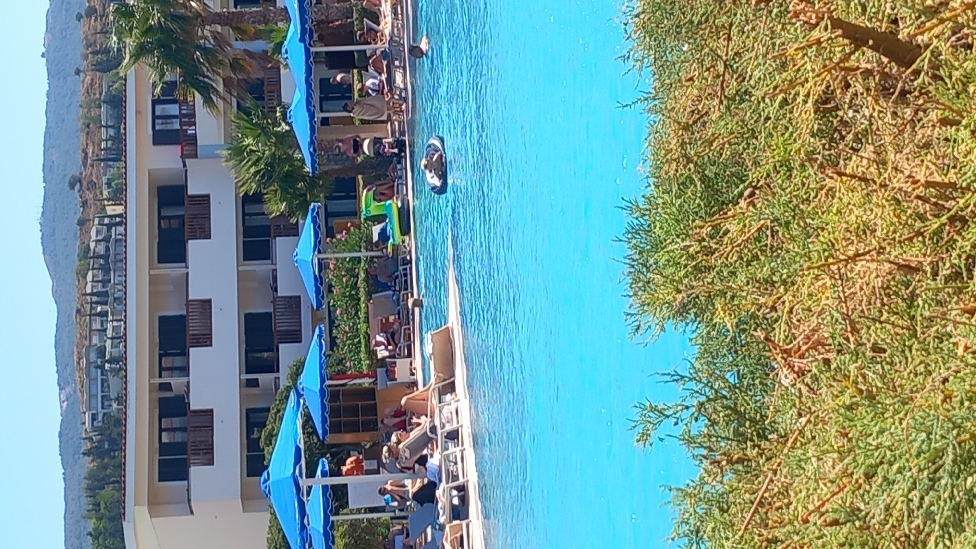 This image shows a resort swimming pool area on the island of Kos, Greece. The pool is bright blue, and several blue umbrellas are set up along the edge. People are visible lounging on chairs and swimming in the pool. In the background, a multi-story hotel building with balconies is visible, and behind that, hills covered with trees and some structures. The foreground is partially obscured by green foliage.
