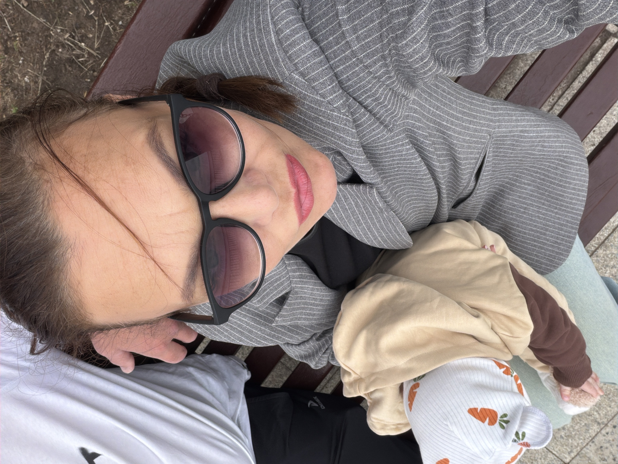 The image is a high-angle, close-up shot of a woman lying on a wooden park bench. She is wearing a gray pinstripe blazer and large black-rimmed sunglasses. Her lips are painted a bright pink. To her right, a baby is visible, swaddled in a tan and brown hooded jacket and wearing a white hat with a carrot pattern. The background shows the textured surface of the park bench and the ground, which appears to be dirt and dry grass. The lighting suggests it is daytime, with natural light illuminating the scene. The woman's head is tilted back, and her eyes are hidden behind her sunglasses, giving a relaxed and possibly content expression. The presence of a park bench and the outdoor setting indicates the scene takes place in a public outdoor space, likely a park. There is no visible text in the image.