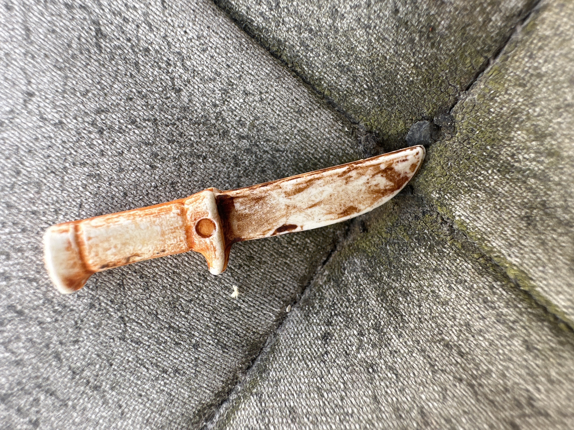 A small, tarnished, toy knife with an orange and white handle and a blade with brown markings lies diagonally on a textured gray surface. The surface appears to be some form of outdoor paving, possibly interlocking stones or a textured concrete slab, with visible seams and areas of green growth, suggestive of moss or algae. The lighting indicates an outdoor setting, with a slight shadow cast by the knife. The image is cropped from a larger scene, providing a close-up view of the object and its immediate surroundings. The location within Lelystad, Netherlands, is not discernible from the visual information alone.