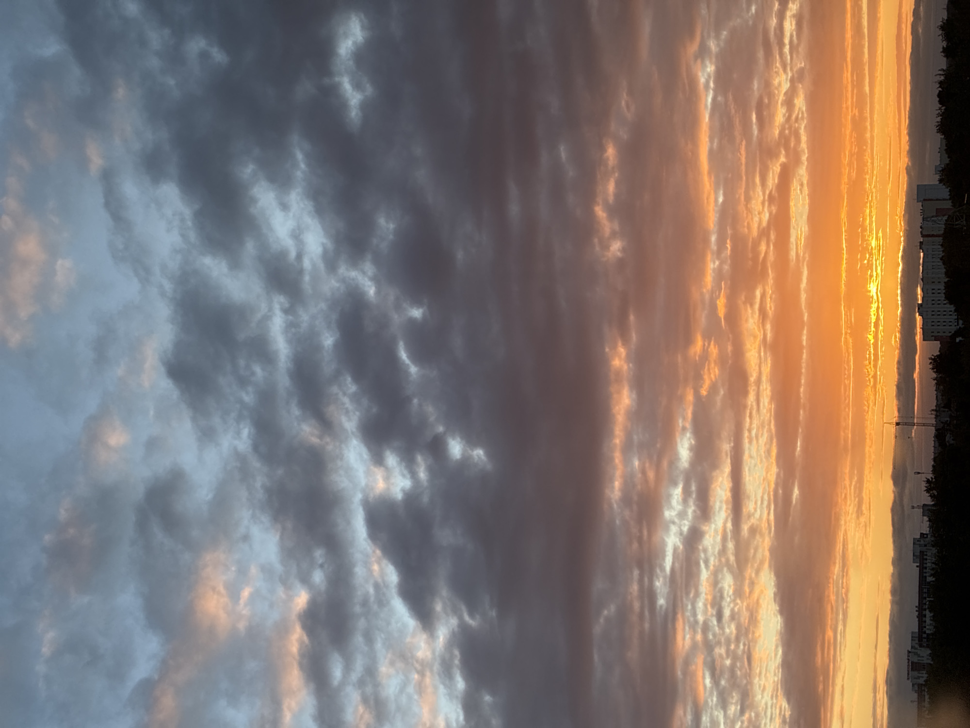 A vertical, wide shot captures a sunset over the cityscape of Unknown, Unknown. The sky is filled with a dramatic display of clouds, transitioning from a pale blue at the top to deep grays and purples, then to vibrant oranges and yellows closer to the horizon. The sun itself is a bright, intensely orange glow peeking through the clouds near the bottom center of the frame. Below the sky, a dark silhouette of trees and buildings forms the lower edge of the image. Various structures, including what appears to be a tall crane, are visible among the buildings, suggesting an urban environment. The overall impression is one of a dramatic and colorful end to the day.