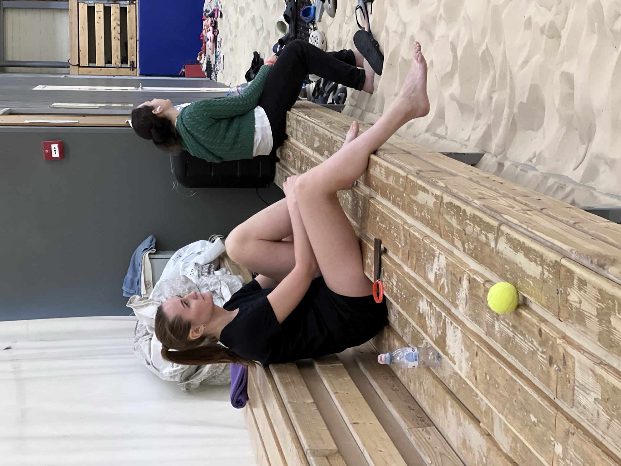 An indoor scene depicts two women resting on a long, light-colored wooden bench within a sandy sports facility in Saint Petersburg, Russia, likely an indoor beach volleyball court. The floor is covered entirely in fine, light-colored sand.
In the foreground, a woman with brown hair tied in a ponytail is seated on the bench, facing right in profile. She wears a black short-sleeved t-shirt and black shorts. Her legs are extended, and she is barefoot, with her left foot slightly suspended above the sand. On the bench beside her are a clear plastic bottle of "Svyatoy Istochnik" brand water, a pair of orange-handled scissors, an orange wristband, and a bright yellow tennis ball. Behind her, a folded purple towel and a pile of mixed light-colored fabrics are also on the bench.
Further down the bench to the right, a second woman is seated, facing left in profile. She wears a green cable-knit hooded sweater over a white t-shirt and dark pants, and has white wireless earbuds in her ears. She is also barefoot, with her feet resting on the sand.
The background features a plain dark grey wall, on which a red fire alarm pull station is mounted. To the right of the second woman, a large black speaker or equipment case is visible. Further right, the wall incorporates wooden slat panels over a blue padded base. Numerous pairs of assorted footwear, including sandals, flip-flops, and clogs, are scattered on the sand in the mid-ground, suggesting they have been removed before entering the sandy area.