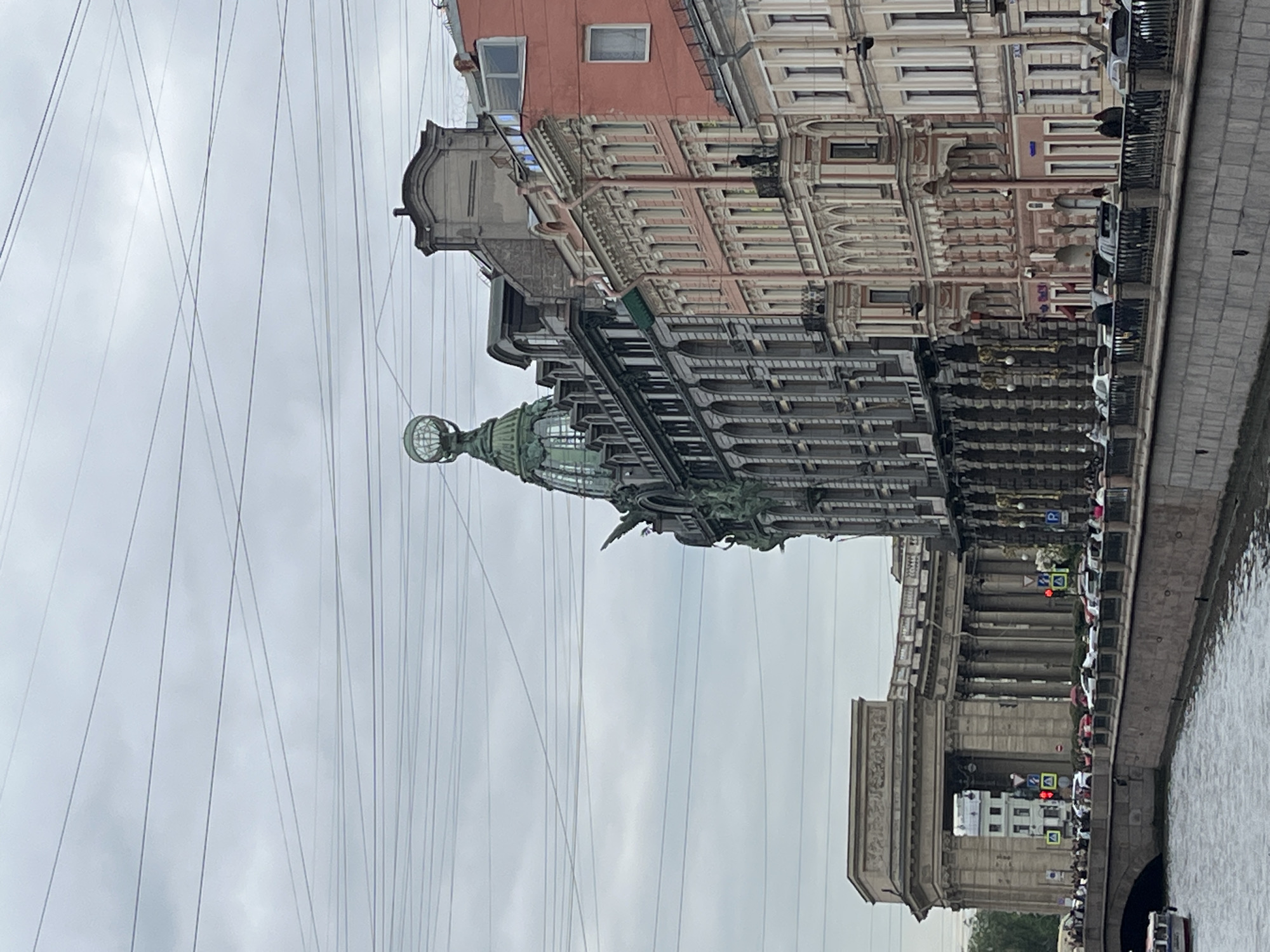 This image showcases the Singer House, also known as the Dom Knigi ("House of Books") building, located in Saint Petersburg, Russia, along Nevsky Prospekt. The photograph, taken at eye level, captures the building's dome with its distinctive glass globe, partially obscured by numerous overhead wires against a cloudy sky. Adjacent to the Singer House, a series of buildings with classic architectural details, including windows and decorative facades, are visible. Below the buildings, there appears to be a bridge or embankment with parked vehicles and pedestrians, as well as a waterway. The composition suggests an urban environment with a mixture of historical architecture and modern infrastructure.