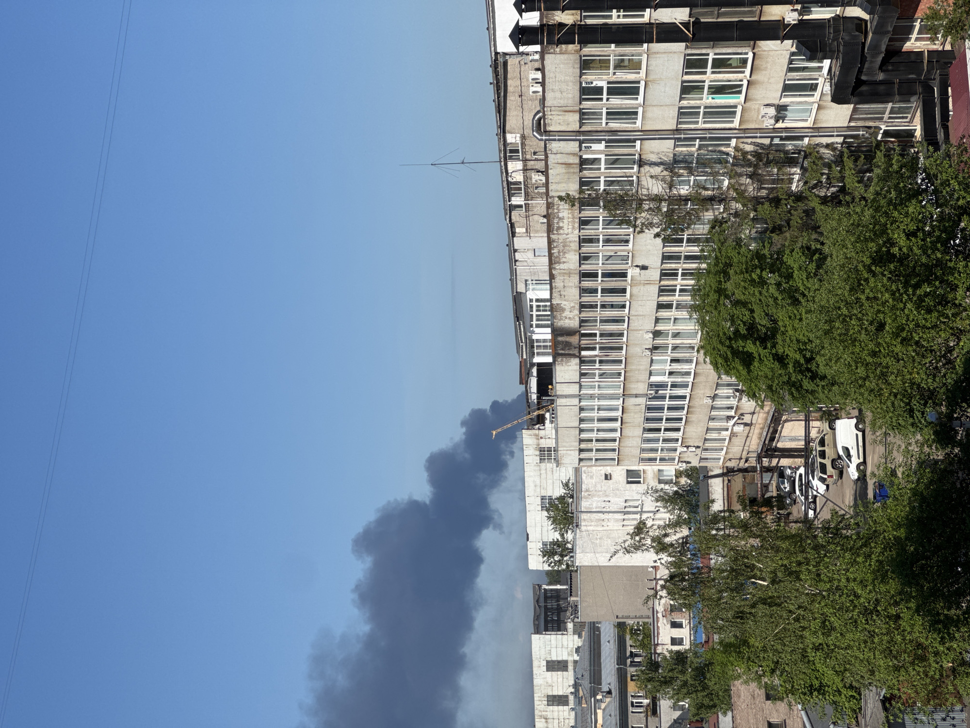 The image captures an urban scene in Saint Petersburg, Russia, under a clear blue sky, depicting a large column of dark grey smoke rising from a mid-ground location.
On the right side of the frame, a tall, multi-story building dominates, characterized by numerous uniform windows and a weathered concrete or light-colored stone facade. Ventilation ducts and pipes are visible along its exterior. Several green, leafy trees are present in the foreground and mid-ground, partially obscuring the lower sections of this building and a small parking area where a few cars, including a white van and a light-colored sedan, are parked.
In the center-left and mid-distance, the most striking feature is the dense, dark smoke plume, indicating a significant fire or industrial event. The smoke billows upwards, starkly contrasting against the otherwise clear, azure sky. Above the smoke, a slender antenna or mast stands atop a distant building, and a faint outline of a crane arm is visible on a rooftop near the smoke's origin. Utility lines or power cables stretch diagonally across the upper left corner of the sky.
The time of day appears to be bright daytime, with ample sunlight. No people are clearly visible, and no discernible text is legible within the frame. The overall atmosphere is one of an active incident due to the prominent smoke.