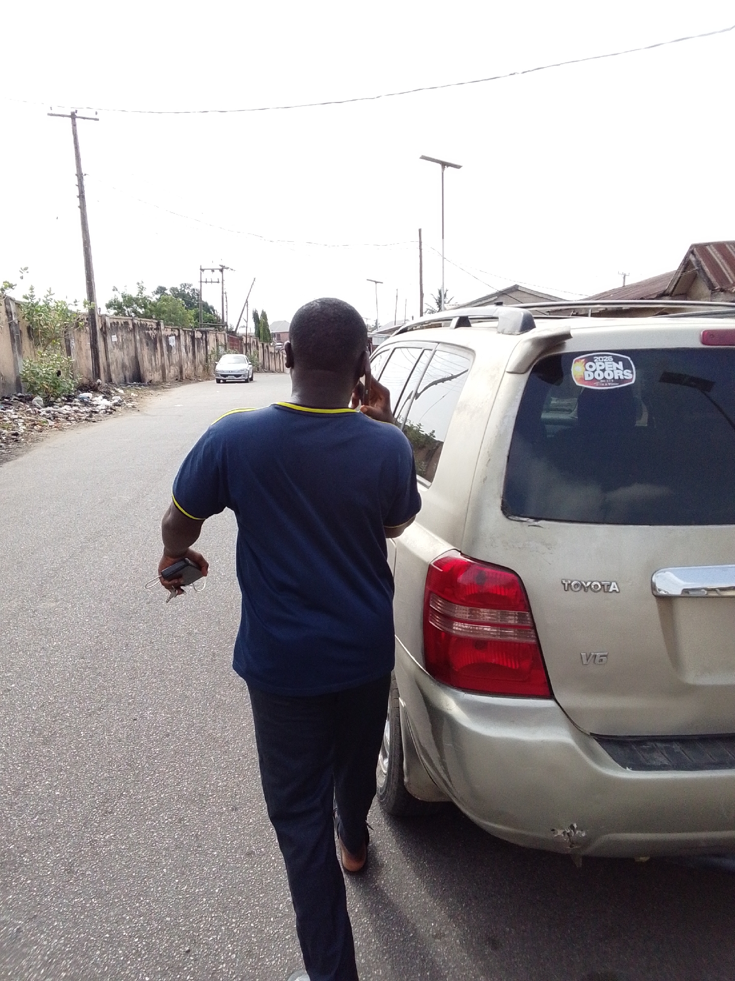 A medium, eye-level shot shows a dark-skinned man walking on an asphalt road beside a beige Toyota Highlander SUV. He wears a navy blue T-shirt with yellow trim and dark trousers and appears to be on a phone call while holding a mobile device and keys. The SUV has a sticker reading "2026 OPEN DOORS" on the rear window and a dent in the rear bumper. The road is bordered by a weathered concrete wall on the left with scattered debris. A light-colored sedan is parked further down the road. Overhead power lines and utility poles are visible in the background against an overcast sky. The location appears to be a residential street in Lokoja, Nigeria.