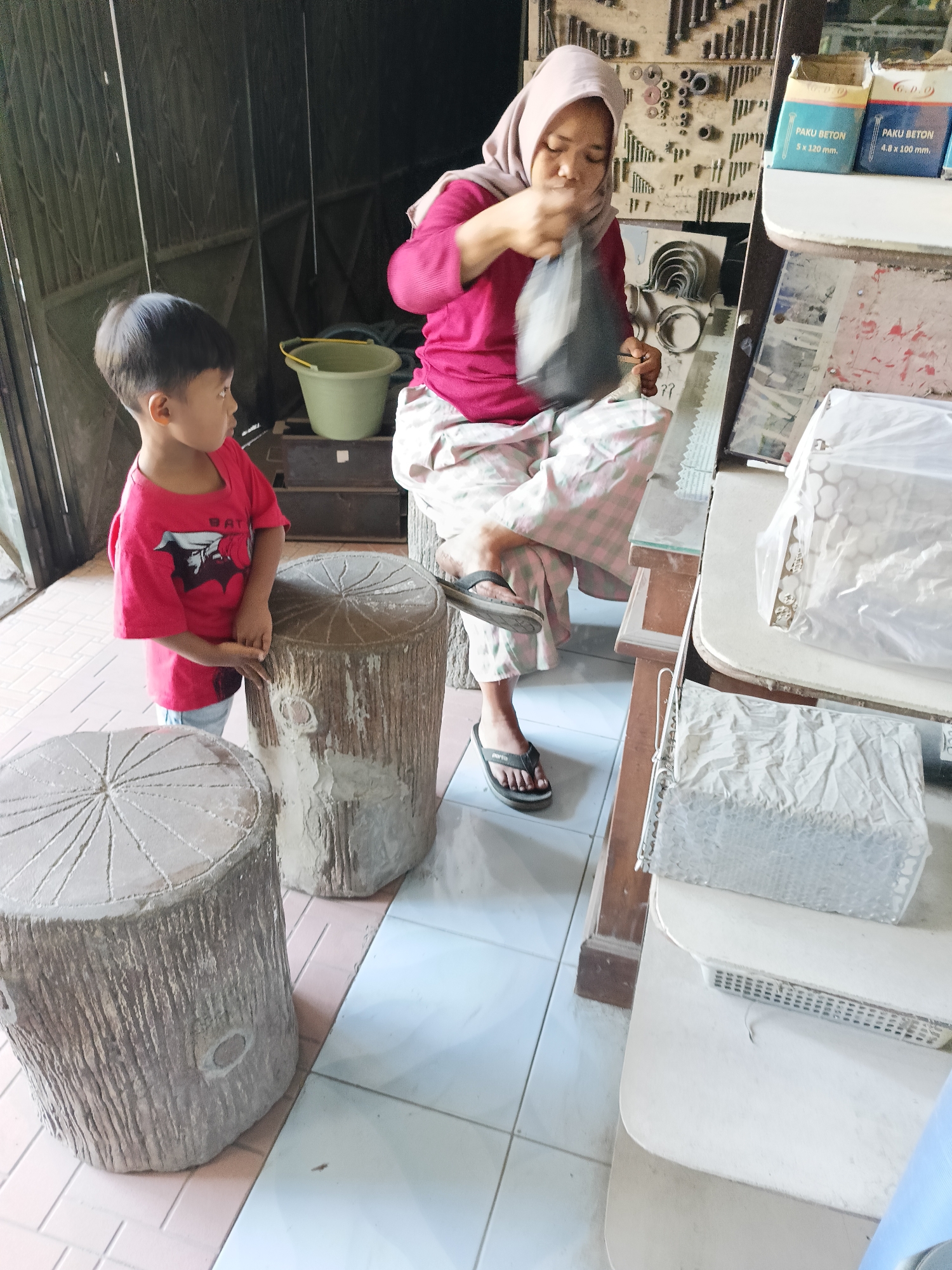 In Sukorini, Indonesia, a woman wearing a pink headscarf and a pink shirt sits on a wooden stool. She is holding a black plastic bag in her hands and appears to be sorting through its contents. The woman is seated in front of a wall adorned with various tools and hardware, possibly in a workshop or storage area. Next to her, a young boy in a red t-shirt stands and looks towards the woman. In the foreground, there are two concrete stools designed to resemble tree stumps. The environment suggests a functional space, perhaps a craftsman's workshop or a retail area for hardware and construction materials, indicated by the bags labeled "PAKU BETON" (concrete nails) on a shelf in the background.