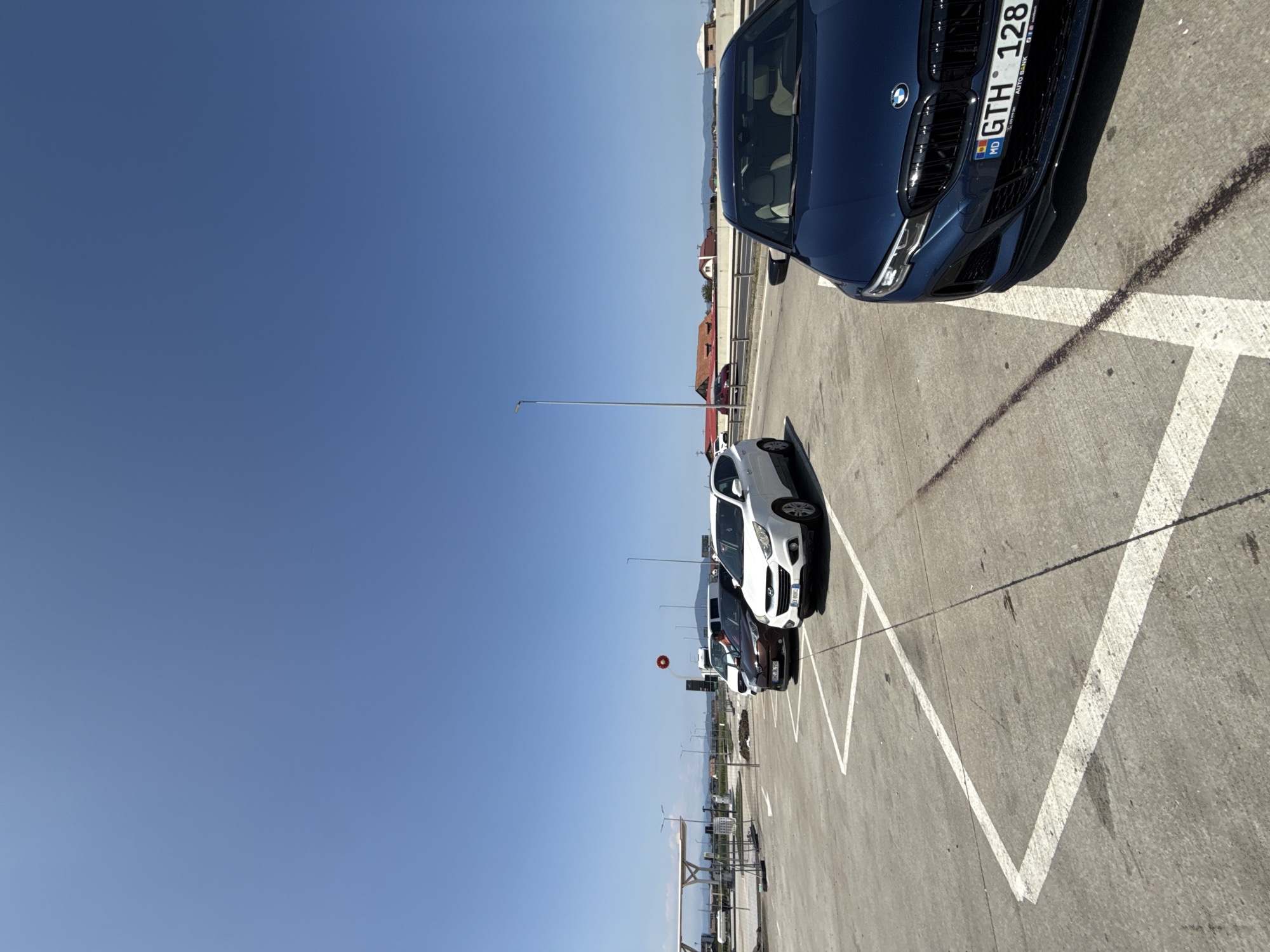The image depicts a parking lot with several vehicles under a clear blue sky. In the foreground, a dark blue BMW is parked, its front grille and headlights clearly visible. Behind it, a white SUV is parked in a designated space marked by white lines on the concrete. Further back, a brown car and a white van are also visible. The parking lot appears to be paved with concrete and has some dirt and debris scattered on the surface. In the background, there is a road with other vehicles, and beyond that, residential buildings with red roofs and some distant hills. A tall lamp post stands in the middle ground. The scene is set in a sunny outdoor environment, likely during the daytime. The location is identified as Sanpetru, Romania.