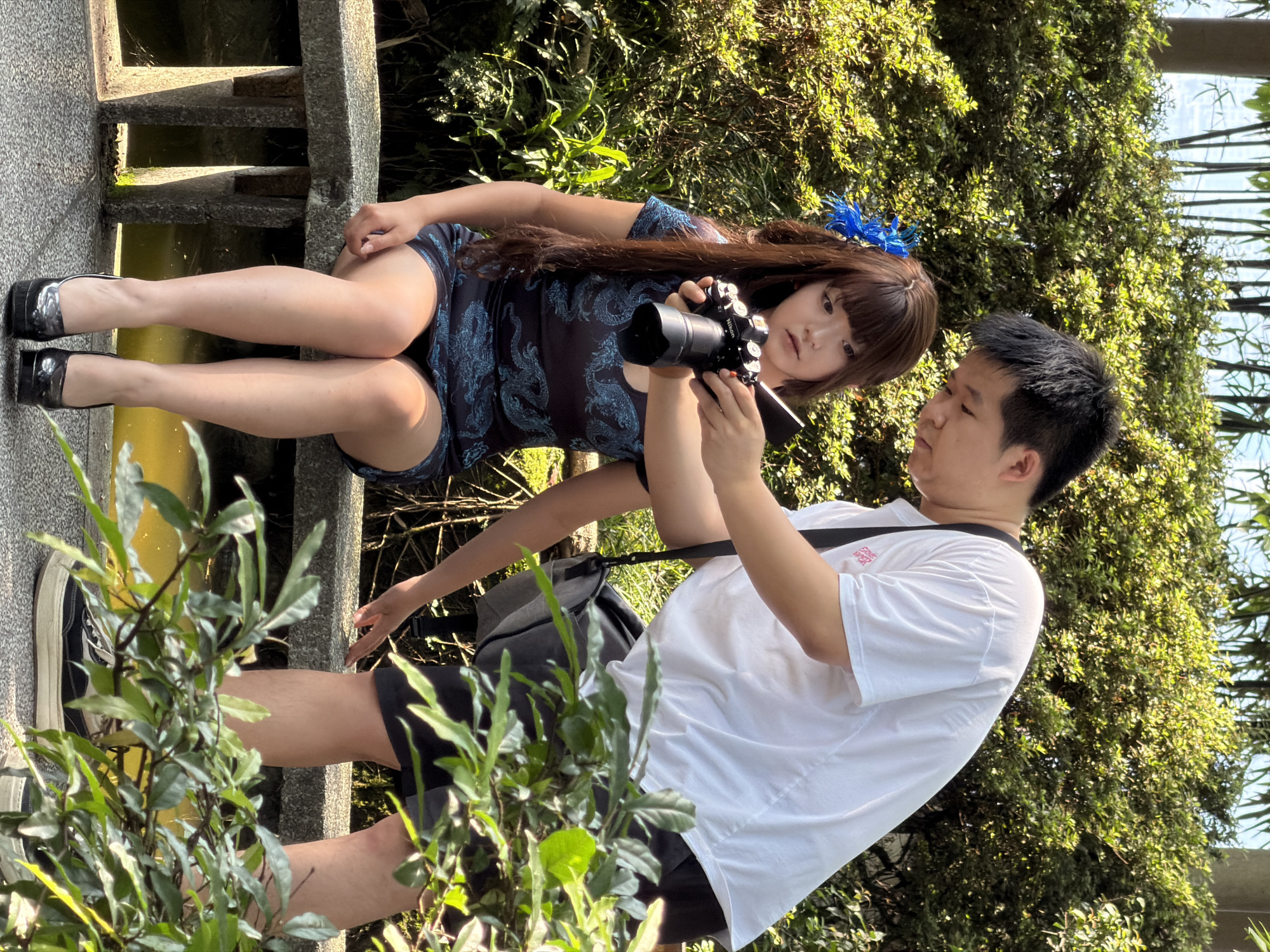The image shows two people in an outdoor setting, possibly a park or garden, in Yuzhong District, China. A young woman is seated on a stone ledge, wearing a dark, patterned dress and high-heeled shoes. She has long, dark hair with a blue decoration on the side and is holding a camera, looking towards the viewer. Next to her, a young man is standing and appears to be taking a photo of her. He is wearing a white t-shirt and dark shorts, with a black strap across his shoulder. Lush green foliage surrounds them, suggesting a natural environment. The lighting indicates it is daytime, with sunlight filtering through the leaves. The scene captures a moment of photography or a photoshoot.
