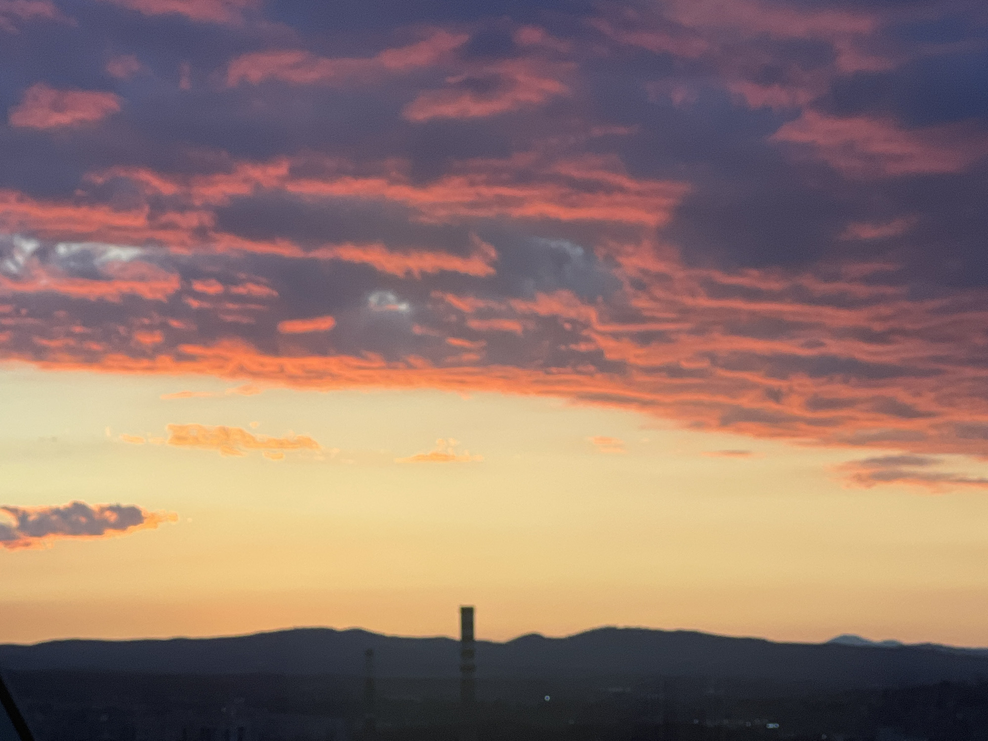 The image captures a vibrant sunset over the city of Sofia, Bulgaria. The sky is filled with layers of clouds, ranging from orange and pink to deep purple, reflecting the setting sun. The horizon line is punctuated by a silhouette of a mountain range and a tall, slender tower, possibly a communication or industrial structure. The lower part of the image suggests an urban landscape, although details are obscured by the distance and dusk. No people are visible in the frame.