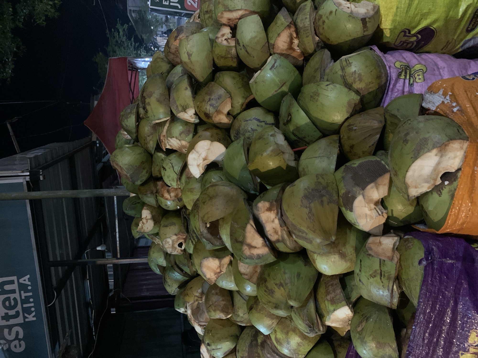 The image captures a nighttime scene at a street vendor stall in Jungke, Indonesia, dominated by a large, conical pile of several hundred green coconuts. Many of the coconuts are partially husked or cut open, revealing the white inner flesh or the dark interior of the shell. They are varied in condition, displaying mottled green exteriors with brown marks, and are stacked directly onto the ground and partially contained within open, brightly colored plastic sacks—purple, orange, and lime green—in the foreground.
In the upper left, a white sign with text reading "és ten" above "K.I.T.A" is visible against a dark teal background, positioned under a dark corrugated metal awning supported by metal beams. A single bare incandescent light bulb hangs from this structure, casting light on the scene. To the right, a red canopy or tent is present in the mid-ground, beyond which dark tree foliage is silhouetted against the night sky. Hints of other structures and a partial sign with "JU" and a red "2" are discernable further in the background on the right. No individuals are visible within the frame, but the arrangement of coconuts and the commercial signage indicate an active vending operation.