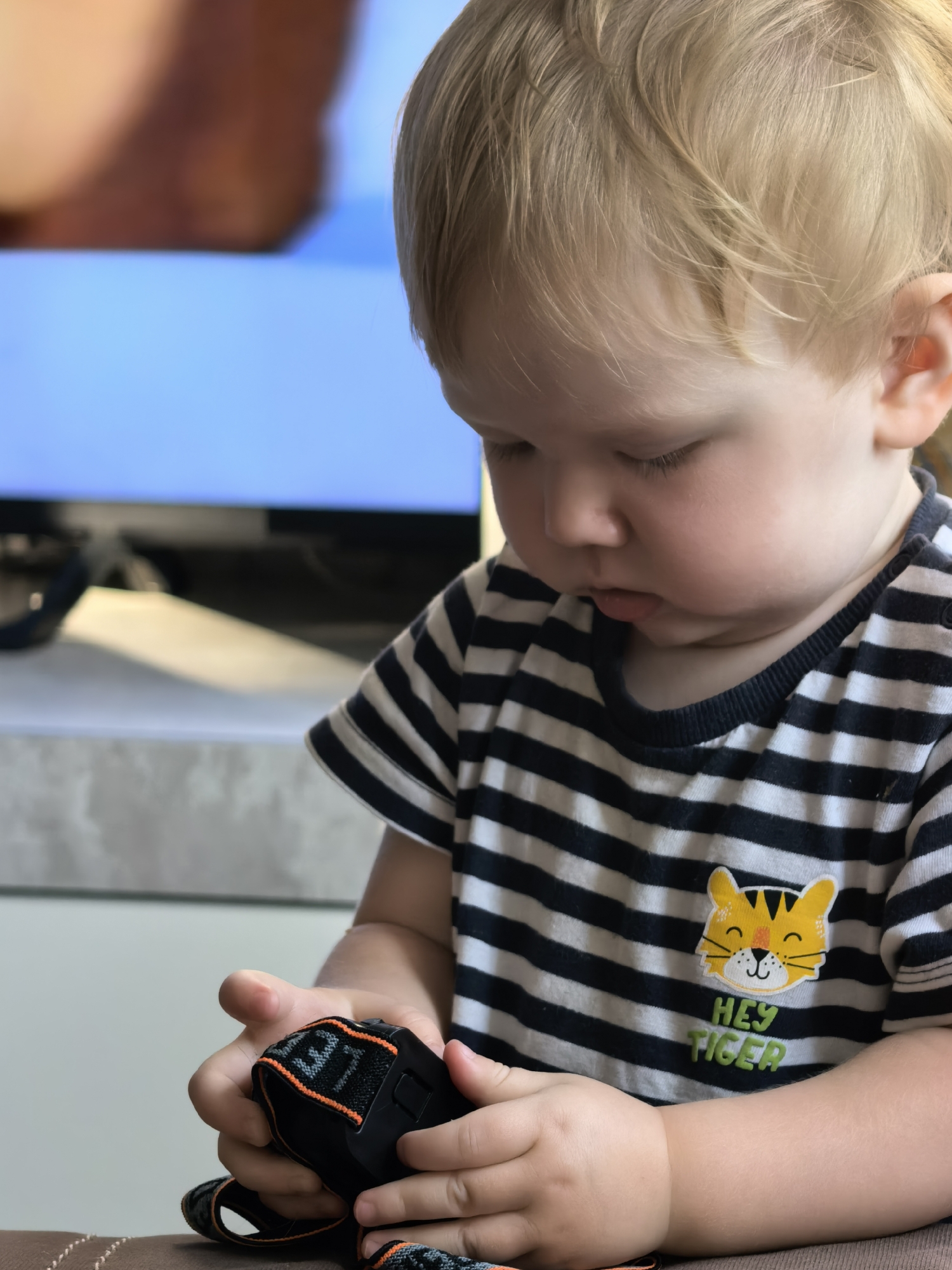 A close-up, medium shot shows a young Caucasian child with blond hair and closed eyes looking down at a black object with orange trim. The child is wearing a black and white striped t-shirt with a yellow tiger face and the words "HEY TIGER" printed on the front. The child's hands are holding the object, which appears to be a camera strap or similar accessory. The background is blurred, showing a light-colored surface and a blue screen, possibly a television. The lighting is soft and natural. The image is cropped to focus on the child and the object. The child's gender and age are not explicitly discernible. The scene is set indoors, likely in a home environment. The city of Sterlitamak, Russia, is not visually represented in the image and cannot be inferred from the provided visual information.