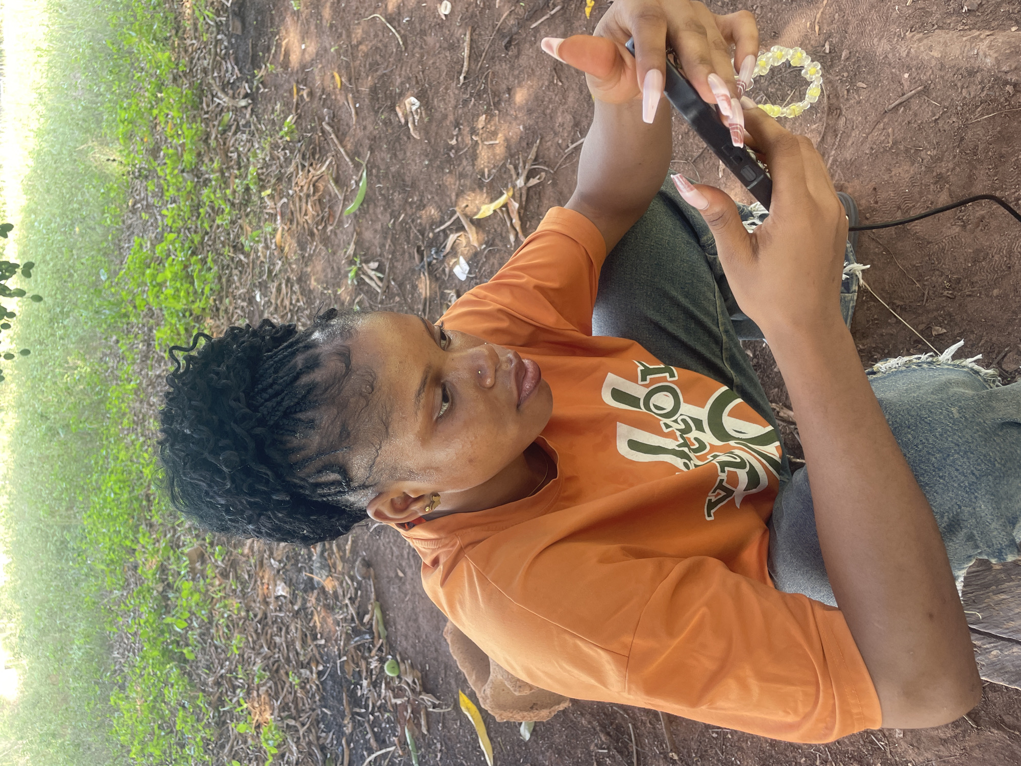 A woman with braided black hair, wearing an orange t-shirt and blue jeans, is sitting on the ground in what appears to be a rural area with grass and dirt. She is holding a black electronic device, possibly a phone, in her hands and looking at it. A black cable is connected to the device. A small, beaded, yellow and white bracelet lies near her. The location appears to be outdoors, possibly in a park or undeveloped area in Ogbe, Nigeria.