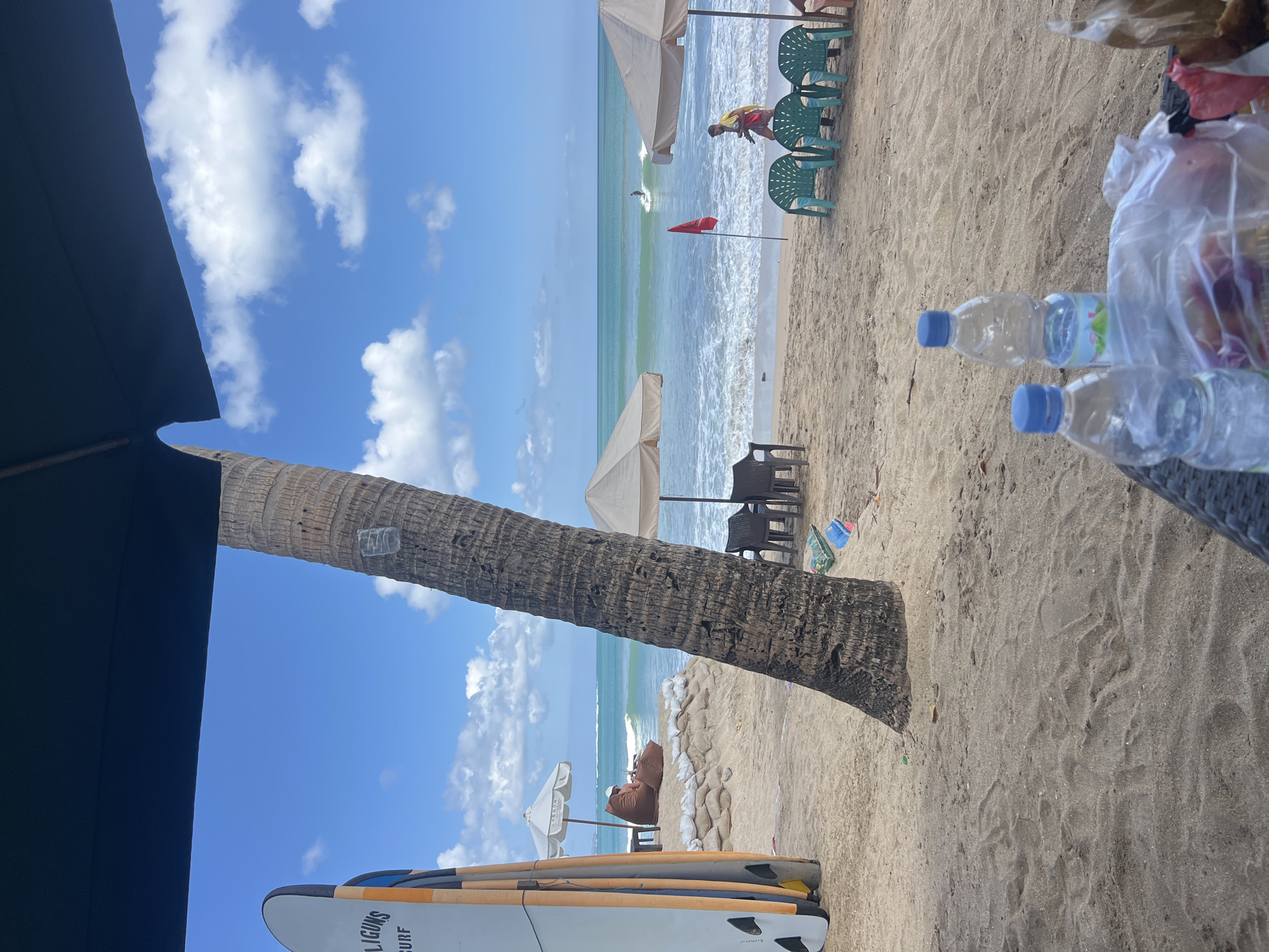 A bright, sunny day unfolds on Kuta Beach, Indonesia, with a clear blue sky punctuated by fluffy white clouds. The foreground is dominated by a sandy beach, with the textured sand showing footprints and ripples. Two plastic water bottles and a bag of food are placed on a woven surface, suggesting a relaxed outing.
Further back, a large palm tree trunk stands prominently, its rough bark creating a natural vertical element. Beyond the tree, beach chairs and umbrellas are set up, offering shade. In the distance, the turquoise ocean waves gently roll onto the shore. A few surfers are visible in the water, one catching a wave. A person in a yellow and red uniform, likely a lifeguard, walks along the beach near a red flag planted in the sand. Several surfboards are propped up on the left side of the frame. The scene evokes a typical tropical beach day in Kuta.