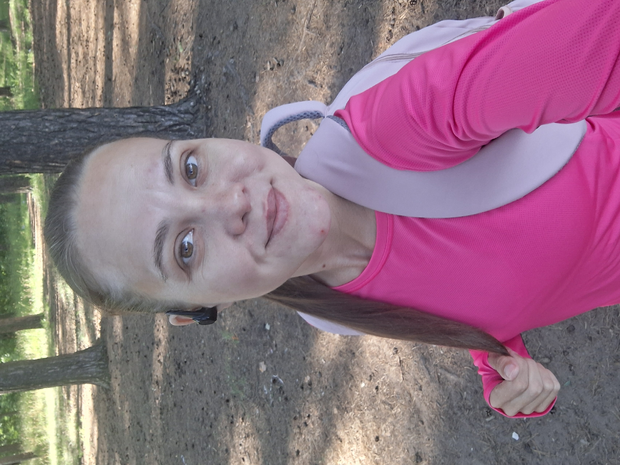 A medium shot shows a woman with brown hair pulled back into a ponytail, wearing a bright pink long-sleeved athletic shirt and a light pink backpack. She has on black wireless earbuds. She is smiling and looking at the camera, holding a strand of her hair with her left hand. The background is a forest floor with pine needles and trees, indicative of a park or wooded area in Chaikovsky, Russia. The lighting suggests it is daytime.