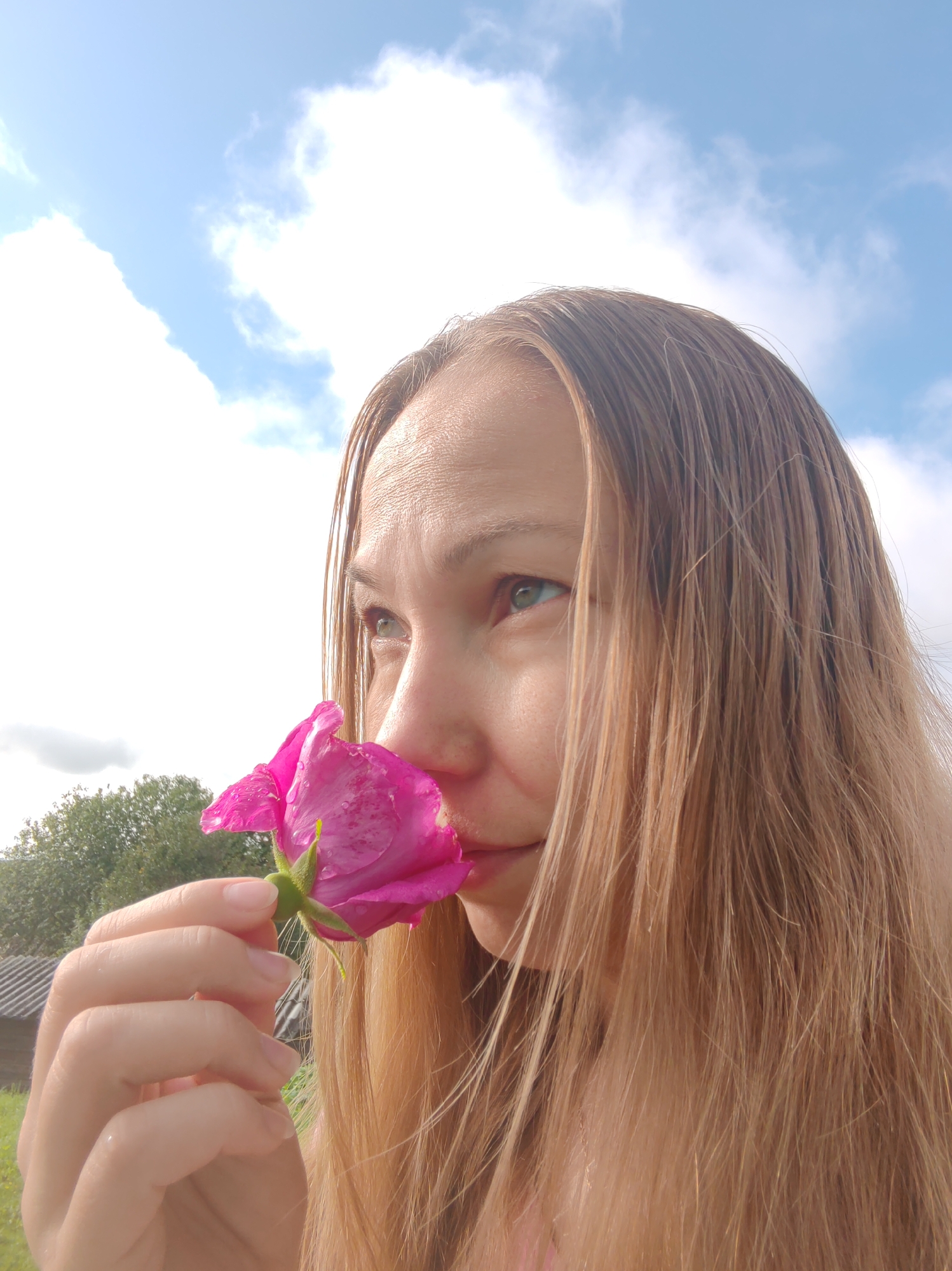 A young woman with long, blonde hair is shown in a close-up profile shot, smelling a pink rose. Her eyes are green and she is looking towards the left side of the frame. The background consists of a bright blue sky with white clouds. In the lower left corner, a portion of a green tree and a rustic building with a dark roof are visible. The overall impression is of a peaceful outdoor scene, possibly in a rural or garden setting. The image does not provide enough information to determine if it is located in the city of Чухлэм, Russia.