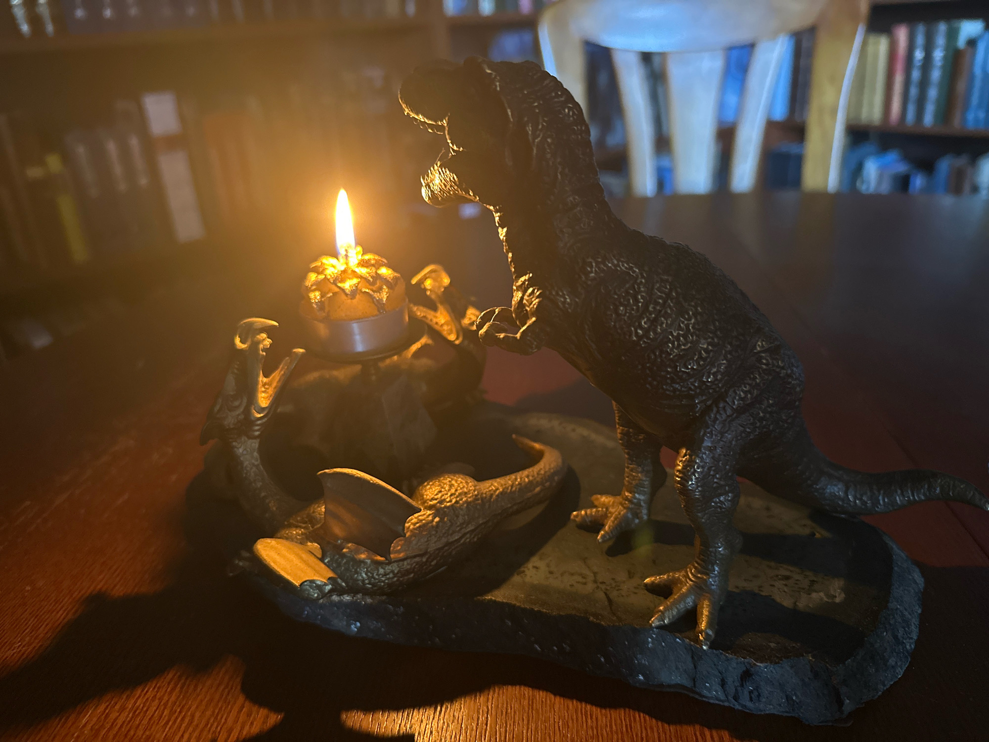 The image shows a close-up, low-angle shot of a decorative candle holder shaped like a prehistoric scene. The candle holder features several dinosaur figures. A large Tyrannosaurus Rex-like dinosaur is prominently positioned on the right side, facing left. In front of it, on a dark, rocky base, are two smaller dinosaur figures. One appears to be a Velociraptor or similar raptor, positioned on the left and looking upwards towards the candle. Another dinosaur, possibly a Pterodactyl with its wings spread, is positioned in the center, its head angled towards the candle. A lit tea light candle burns brightly in a decorative golden holder atop a rocky mound in the center of the scene. The background is out of focus, showing what appears to be bookshelves filled with books and the out-of-focus silhouette of a chair. The lighting is dim and atmospheric, with the candle flame casting a warm glow and long shadows on the wooden surface. This scene is located in a room within the city of Murmansk, Russia.
