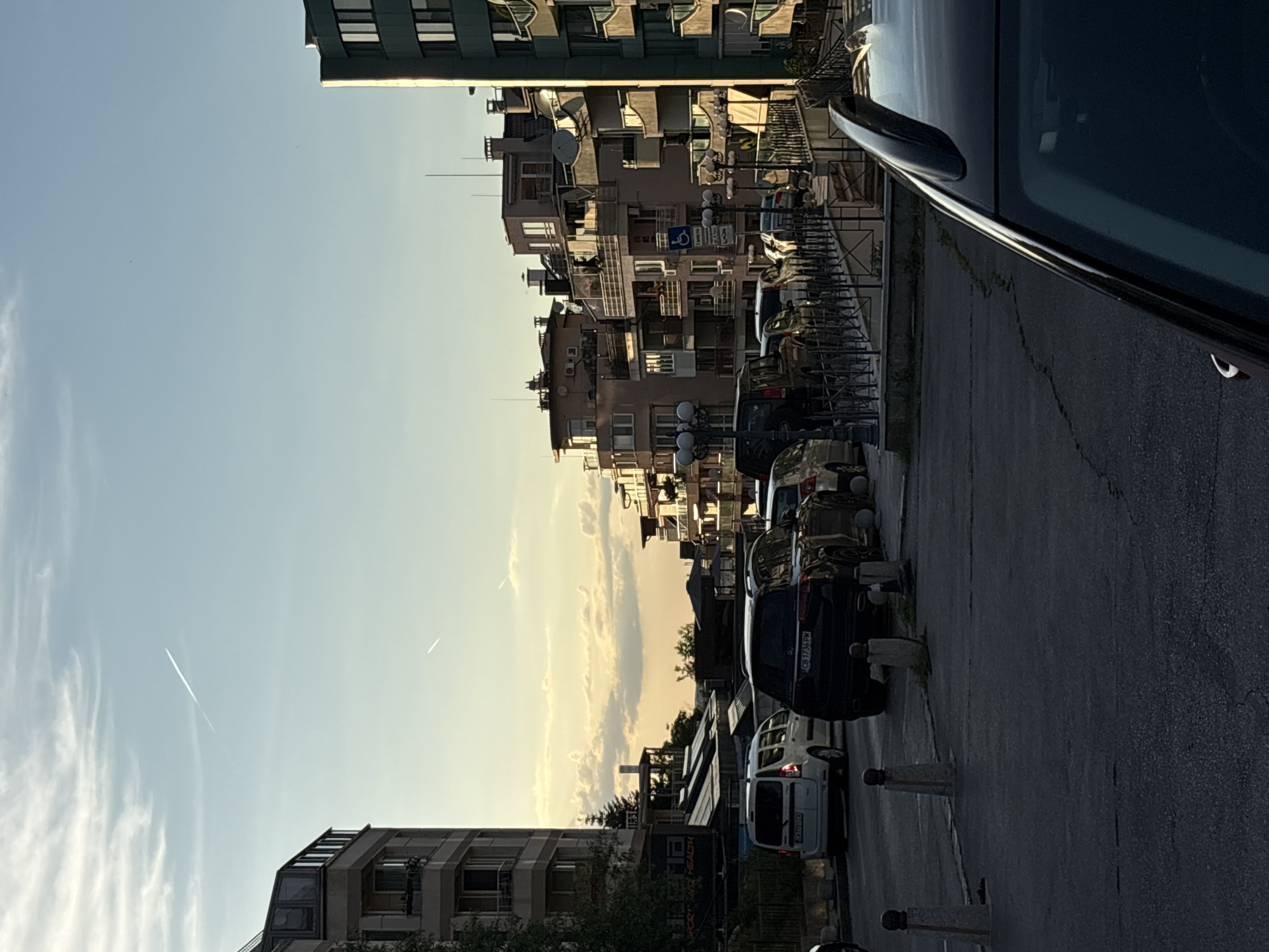 The image captures a street scene in Sofia, Bulgaria, likely during the golden hour as indicated by the warm lighting. A multi-story apartment building is visible on the left, with balconies and windows. To the right, a row of apartment buildings with balconies, satellite dishes, and architectural details line the street. Several cars are parked along the road, including a white van and a black SUV. Streetlights and bollards are present on the sidewalk. The sky is a clear blue with some wispy clouds and two faint contrails from airplanes. The overall impression is of a residential urban environment bathed in soft evening light.