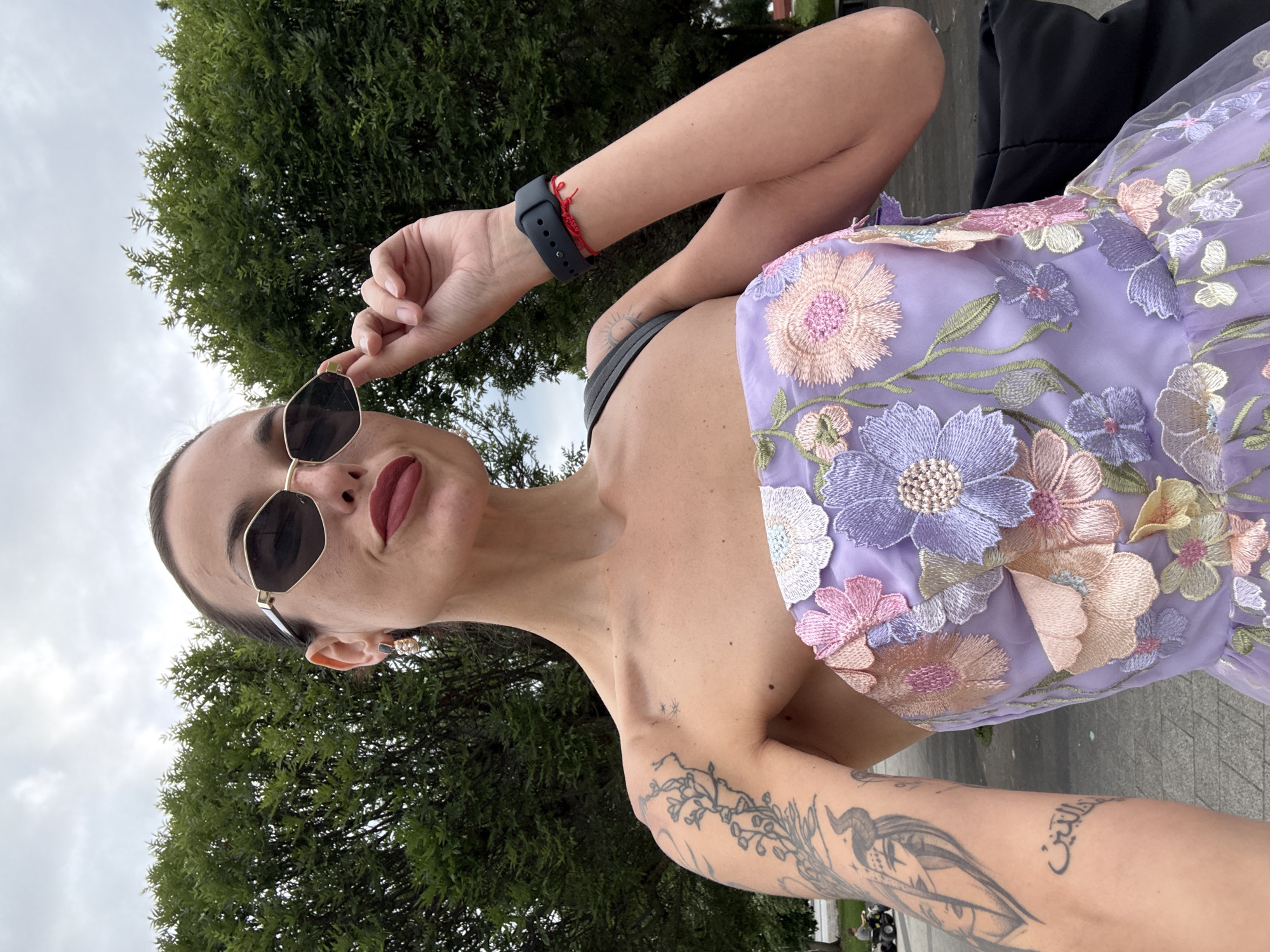 A woman with dark hair pulled back and a pale complexion poses for a selfie outdoors. She wears large, angular sunglasses, dark red lipstick, and a strapless lavender dress adorned with colorful floral embroidery. She has tattoos on her left arm and shoulder, and is wearing a dark smartwatch and a red string bracelet on her right wrist. The background features lush green trees and a cloudy sky. In the far background, a person is seen on a small all-terrain vehicle. The woman is standing on a paved area, suggesting an urban or park setting. The location is identified as Pleven, Bulgaria.