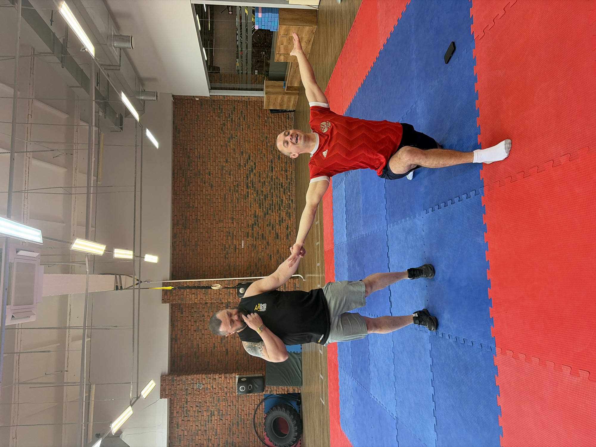 Two men are engaged in an activity within a gym setting in Chaikovsky, Russia. One man, positioned on a blue and red mat, wears a red shirt, black shorts, and one white sock; he is extending his arm upward. The other man, standing on the same mat, wears a black tank top and gray shorts; he is holding the first man's hand. The background includes exposed brick walls, gym equipment, overhead lighting, and a visible structural ceiling.
