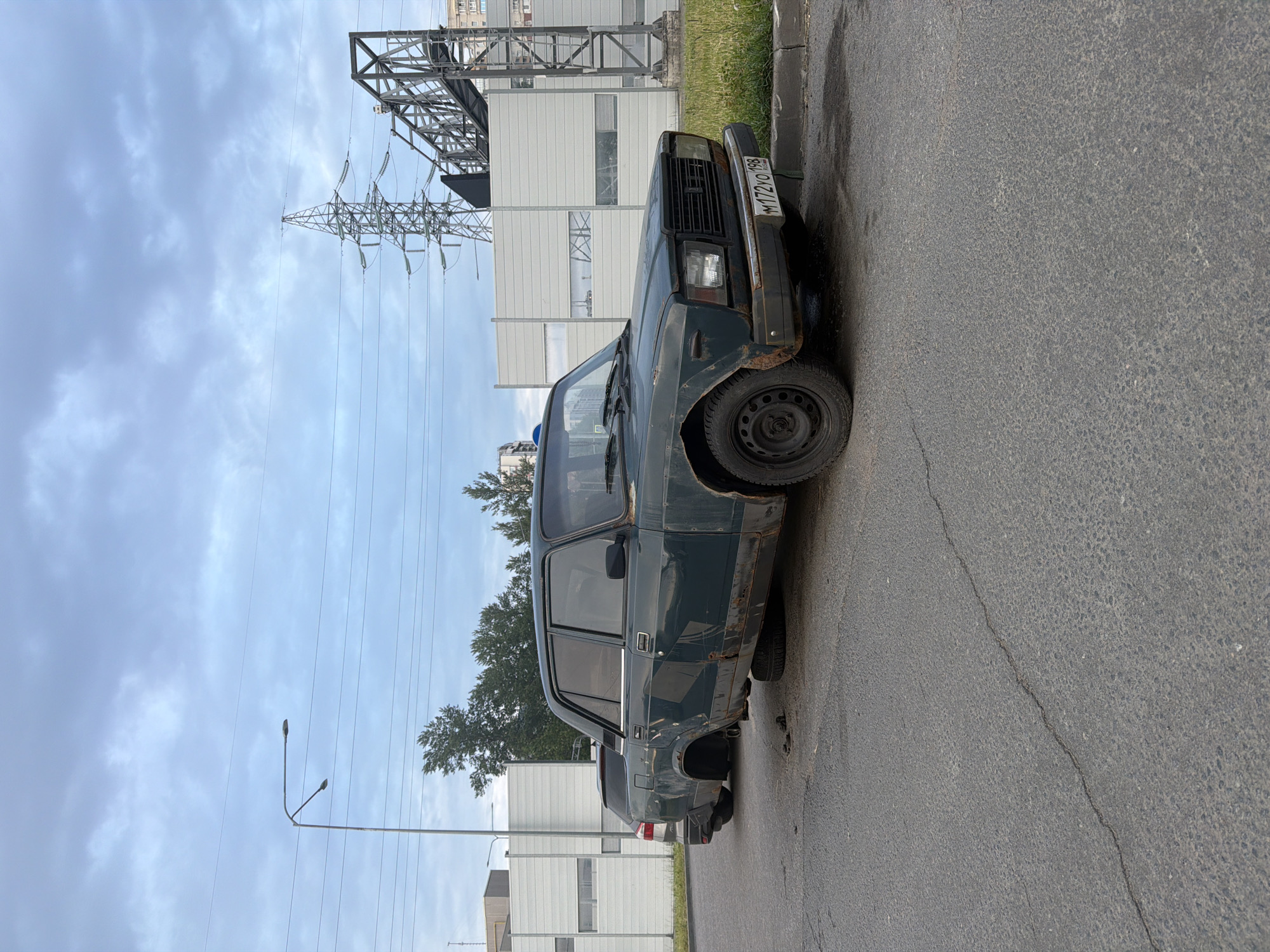 An old, dark green sedan is parked on an asphalt road in a suburban area. The car is in poor condition, with significant rust and damage to the body. One of its rear wheels is missing, and the car is resting on the axle. Another flat tire is visible underneath the car. The car has a license plate that reads "M172YO 198". In the background, there is a modern white building with large windows, a power line tower, and a street light. The sky is overcast with clouds. The scene appears to be an abandoned or neglected area.