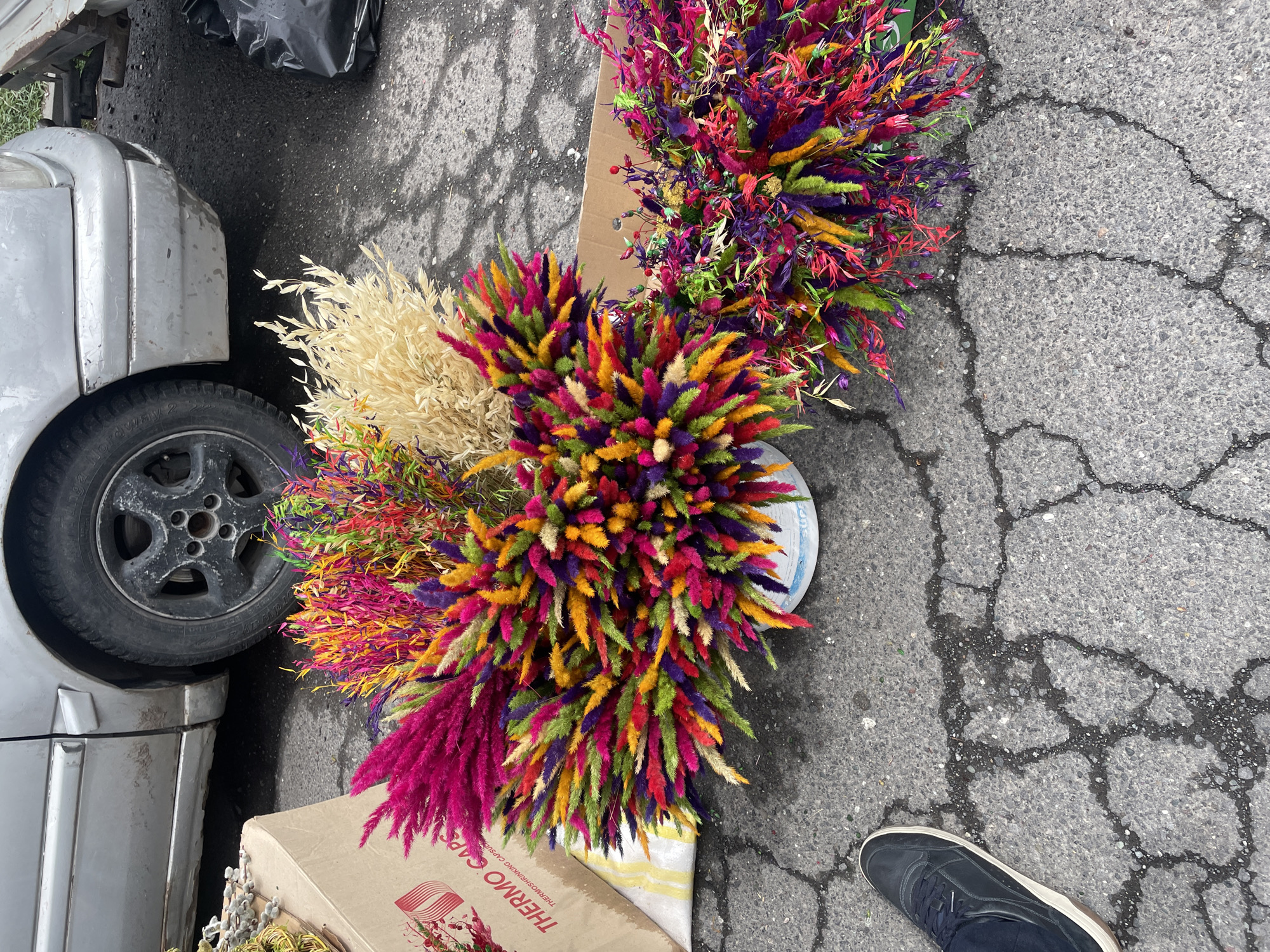 Observed from an overhead perspective, the scene is set on a cracked asphalt surface, presumably a street, adjacent to a silver car and a black plastic bag, with a portion of a shoe visible in the lower right corner. Dominating the composition are three vividly colored floral arrangements; one is a pale beige, and the other two are a vibrant mix of reds, purples, yellows, and greens, positioned on and around a cardboard box with the words "THERMO SHRINKING CAPSULES" visible on its side. The environment appears to be an urban area, specifically, the city of Yerevan, Armenia. No people are fully visible, only a foot and part of a leg indicating human presence. The overall atmosphere suggests a market or a roadside sale of floral items in Yerevan.