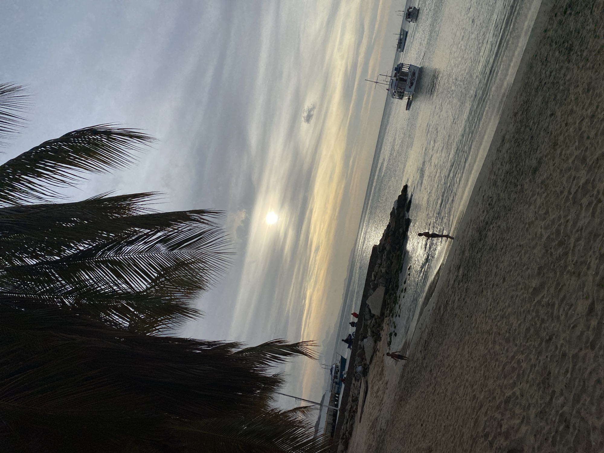 The image depicts a coastal scene in Juan Griego, Venezuela, at either sunrise or sunset, indicated by the low sun visible through a partially overcast sky. The foreground on the left is dominated by the dark silhouettes of palm fronds extending from the top and left edges. A sandy beach occupies the lower left portion of the frame. Two individuals, appearing to be children or youths, are present on the beach; one is walking near the water's edge, and another stands closer to the water, both largely silhouetted. To their right, a rocky barrier or sea wall separates the sand from a concrete pier or walkway. On this walkway, a group of approximately four individuals is gathered, and one person is seated on a motorcycle. A blue and white boat is moored alongside the pier. The midground and background consist of calm water with several boats. A prominent white boat with a cabin and a mast-like structure is moored in the mid-right. Two smaller, darker boats are visible further out on the water towards the right horizon. Distant landmasses or hills are faintly visible on the horizon. The sky features layered clouds, predominantly grey with areas illuminated by the sun's warm glow, which casts a bright reflection on the water's surface.