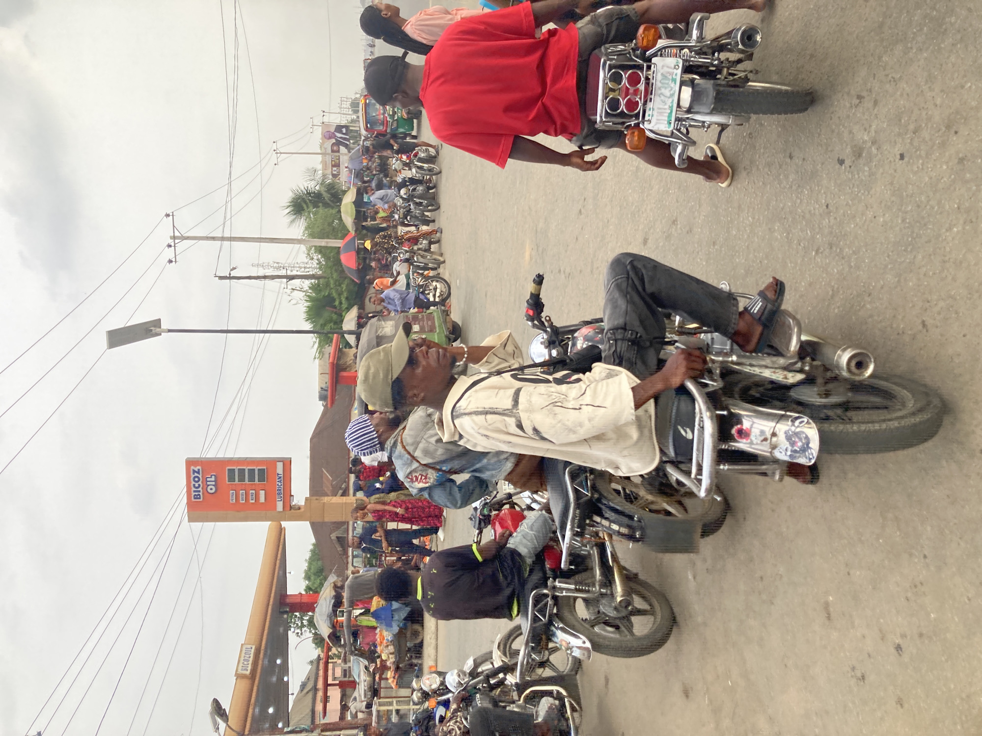 The image depicts a busy street scene in Ura Nta Umuarandu, Nigeria. Numerous motorcycles (okadas) are lined up, many with passengers. The individuals are wearing casual clothing. A man in a red shirt is riding on the back of one motorcycle. The environment appears to be overcast and humid. In the background, there is a BICoz OIL gas station. Power lines are visible overhead. There is a sense of congestion and activity on the street.