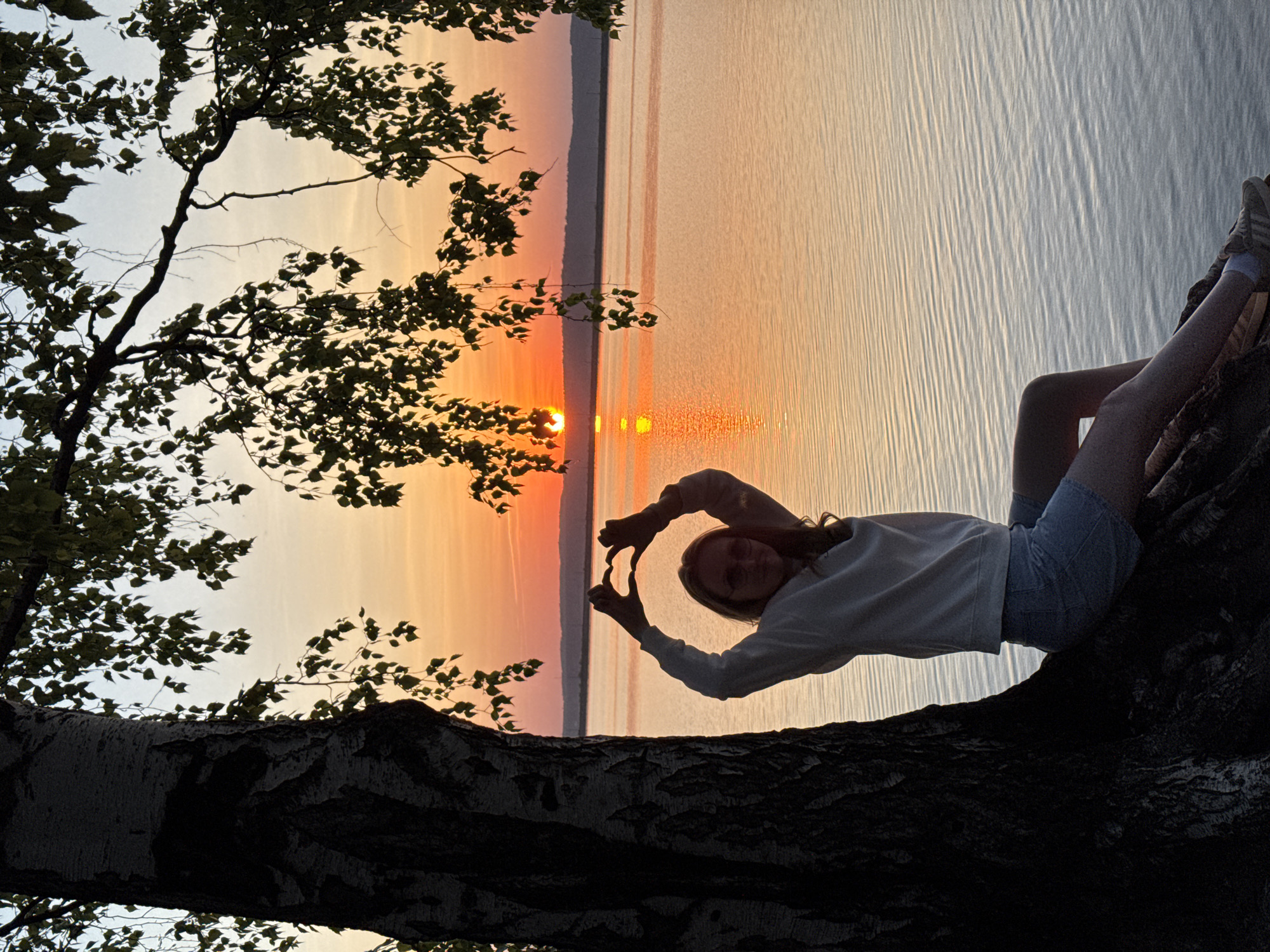 A medium shot shows a woman sitting on a tree branch in front of a sunset over a body of water. The woman is positioned on the right side of the frame, reclining against the tree branch. She is wearing a light-colored long-sleeved shirt and short denim shorts. Her hands are raised, forming a heart shape with her fingers, framing the setting sun. A tree with sparse foliage is silhouetted on the left side of the frame. In the background, the water reflects the orange hues of the sunset, and land is visible across the water. The picture was taken in the city of Chaikovsky, Russia.